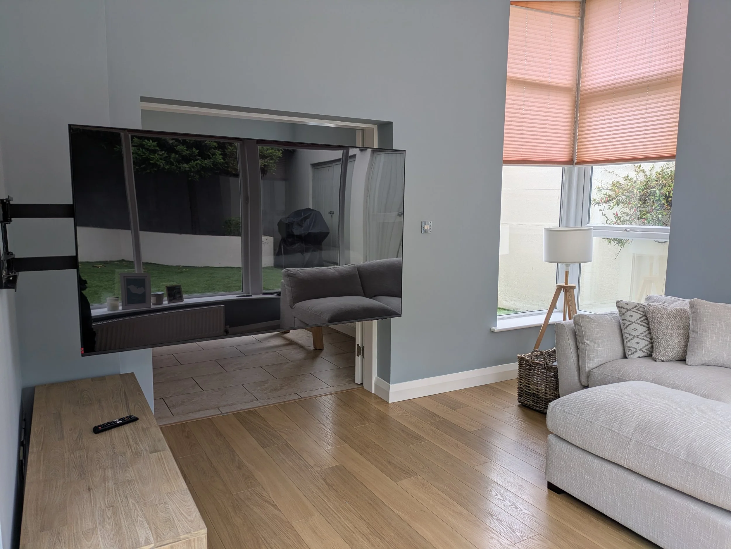 Living room with a beige sofa, wooden floor, and a tall floor lamp near a window with pink blinds. A mounted flat-screen TV is on the wall, facing a small wooden table with a remote control. Through the doorway, an enclosed patio with artificial grass and a barbecue grill is visible.