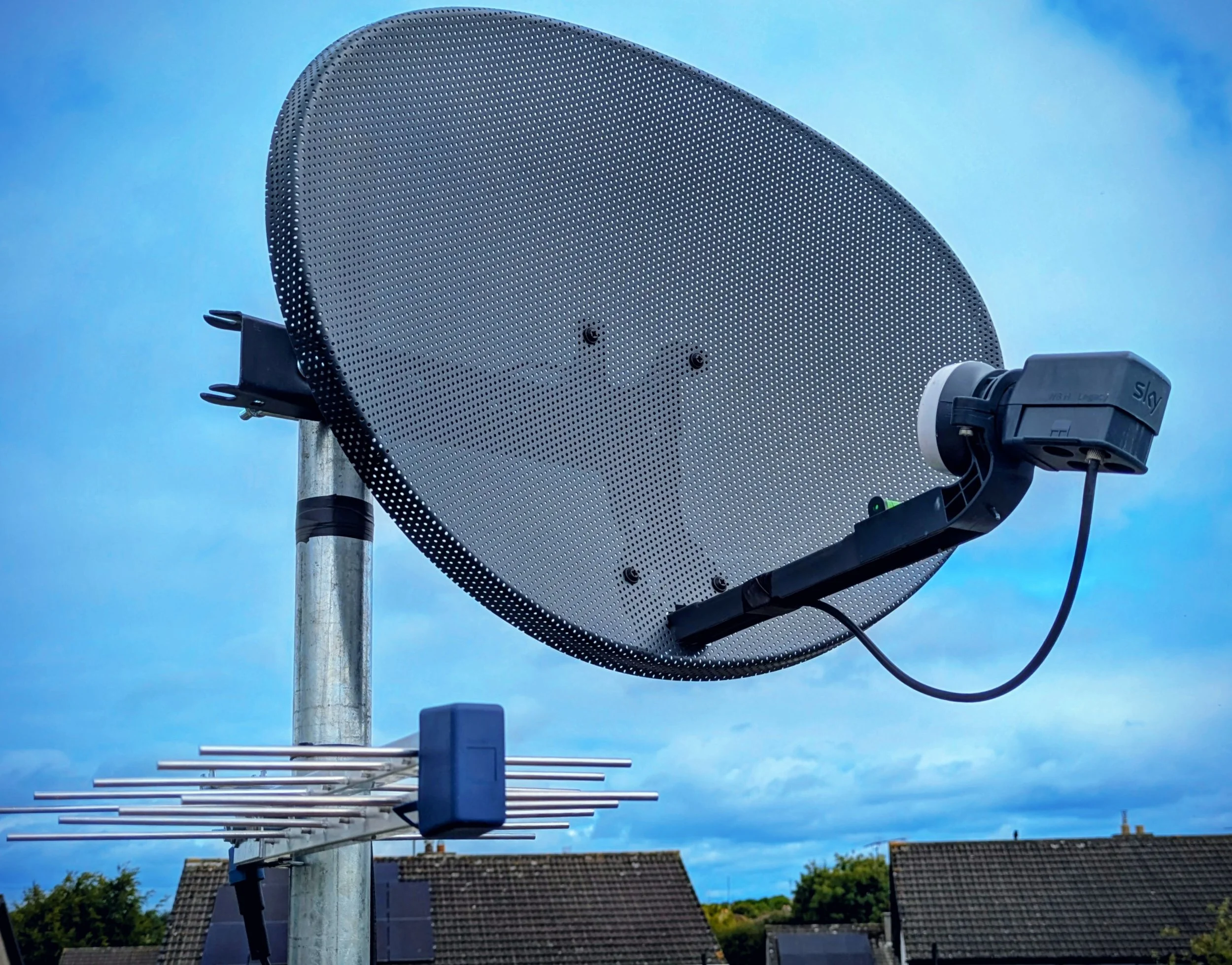 Close-up of a satellite dish mounted on a pole with a smaller antenna below it, against a blue sky with some clouds and rooftops in the background.