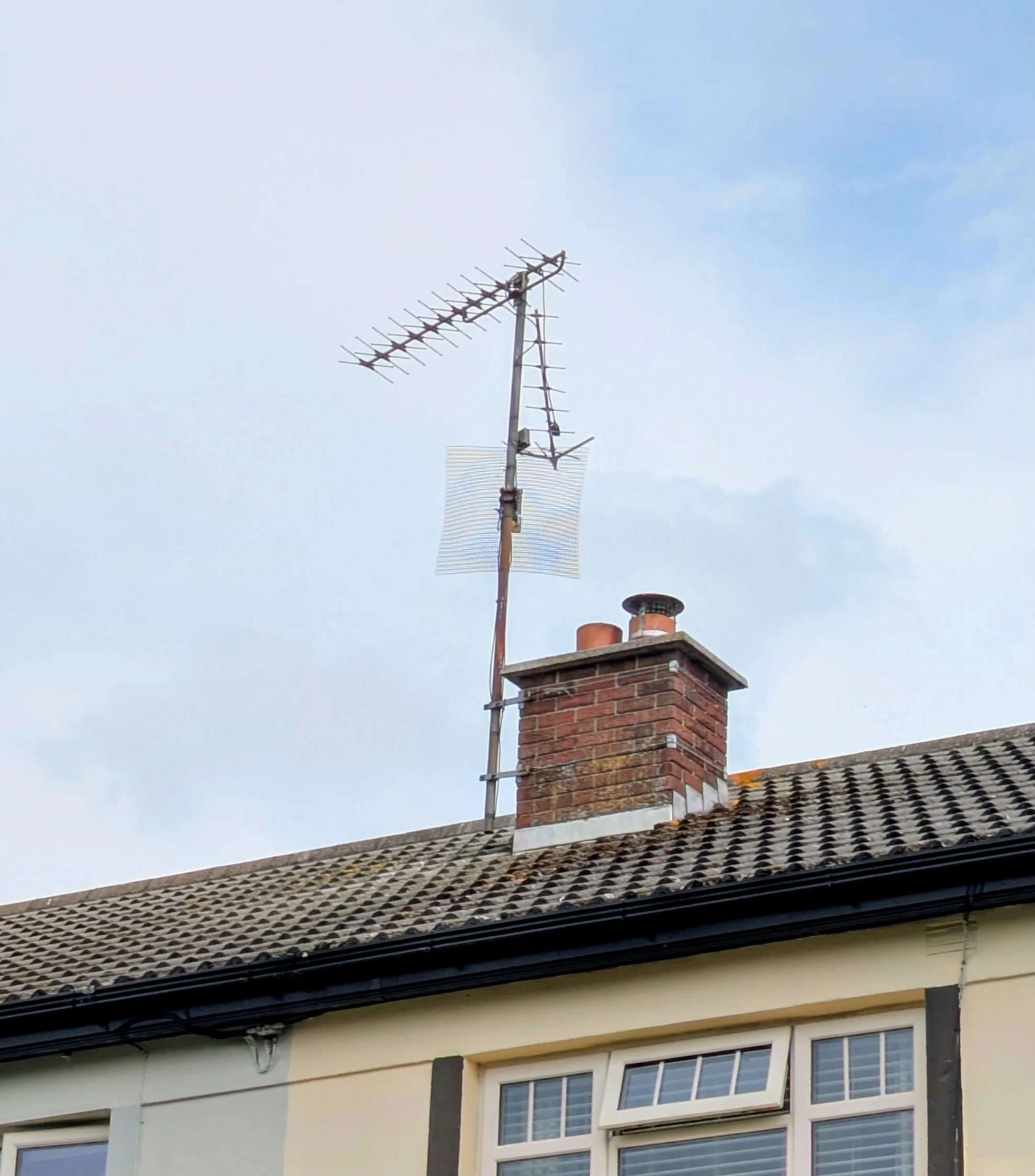 A TV antenna on top of a house roof with a brick chimney and cloudy sky in the background.
