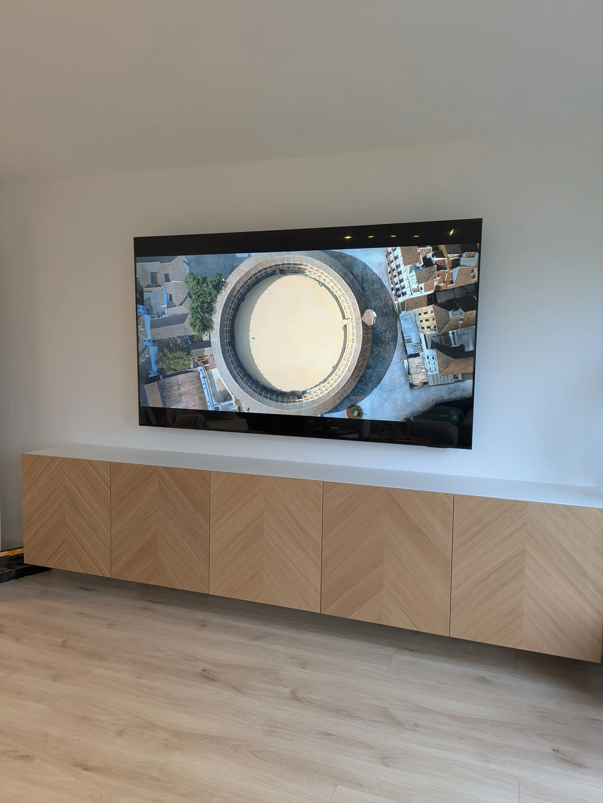 A flat-screen TV mounted on a white wall above a wooden cabinet with four panels in a modern living room. The TV displays an aerial view of a circular building with surrounding rooftops and streets.
