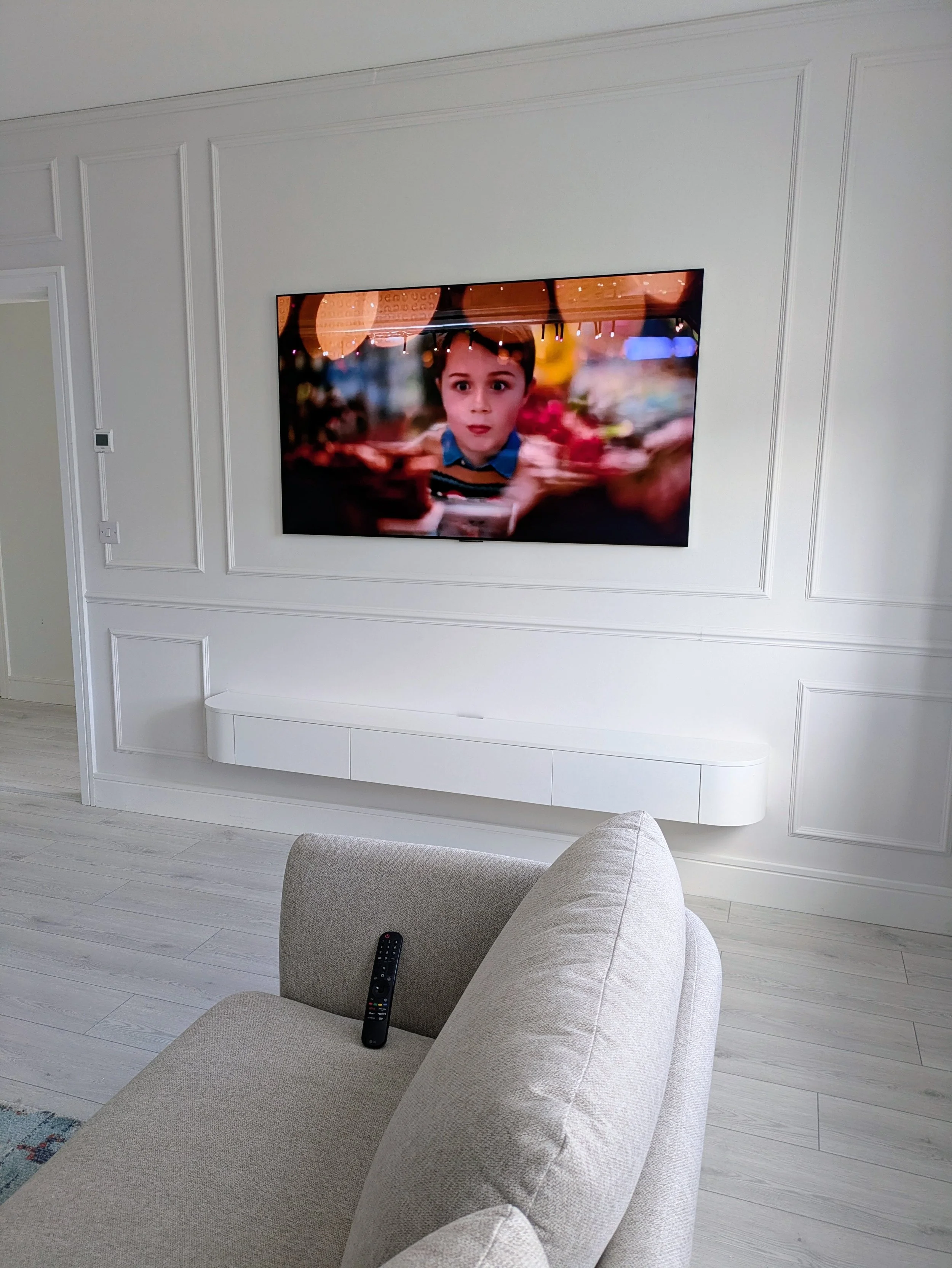 Living room with mounted TV displaying a child's face, beige sofa with remote control on armrest, white wall with decorative paneling, and white wooden flooring.