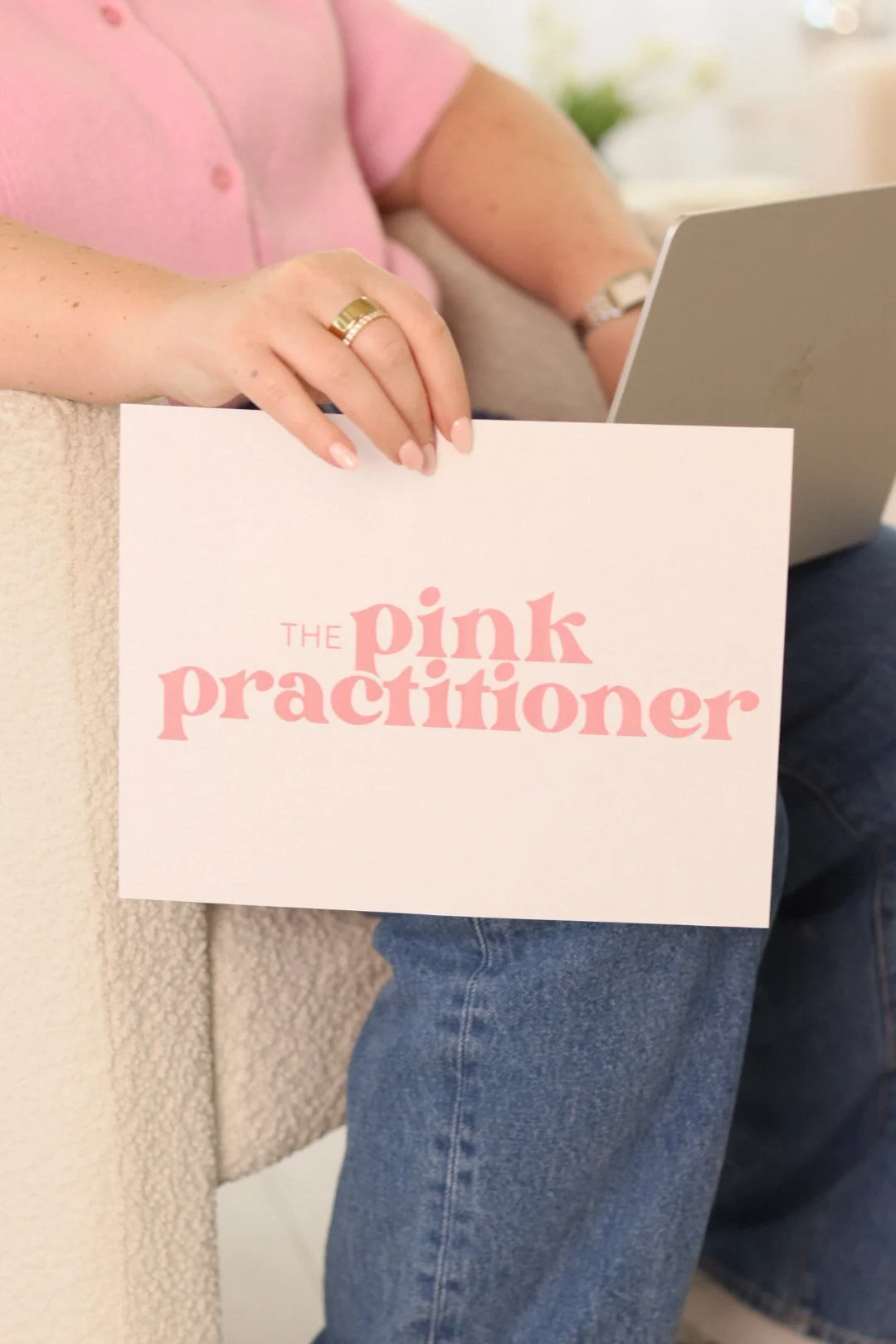 Person holding a card with pink text that says 'The pink practitioner' in front of a laptop, sitting on a beige couch.