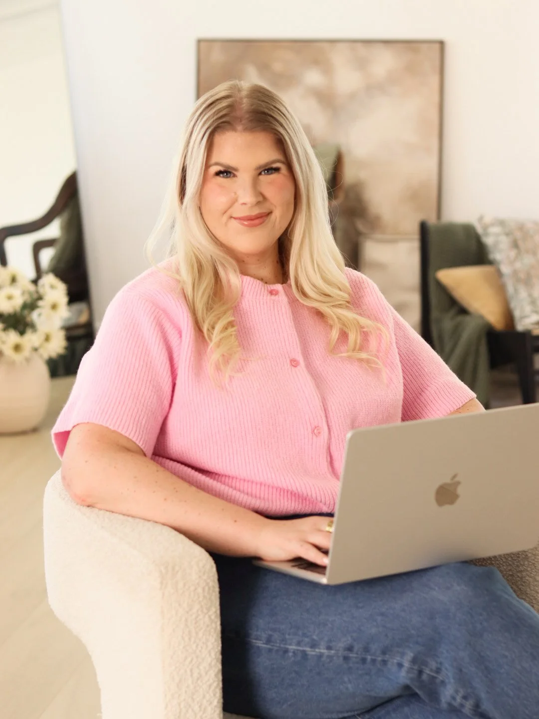 A young woman with long blonde hair and a pink cardigan sitting on a beige chair using a silver MacBook.