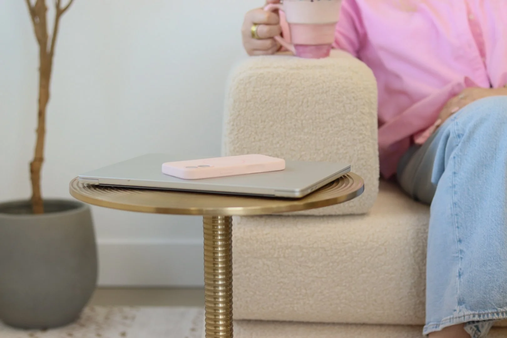 Side view of a beige sofa with a person sitting on it, wearing a pink shirt and blue jeans, holding a pink mug. A small round side table in front of the sofa holds a laptop and a pink phone. A potted plant is in the background.