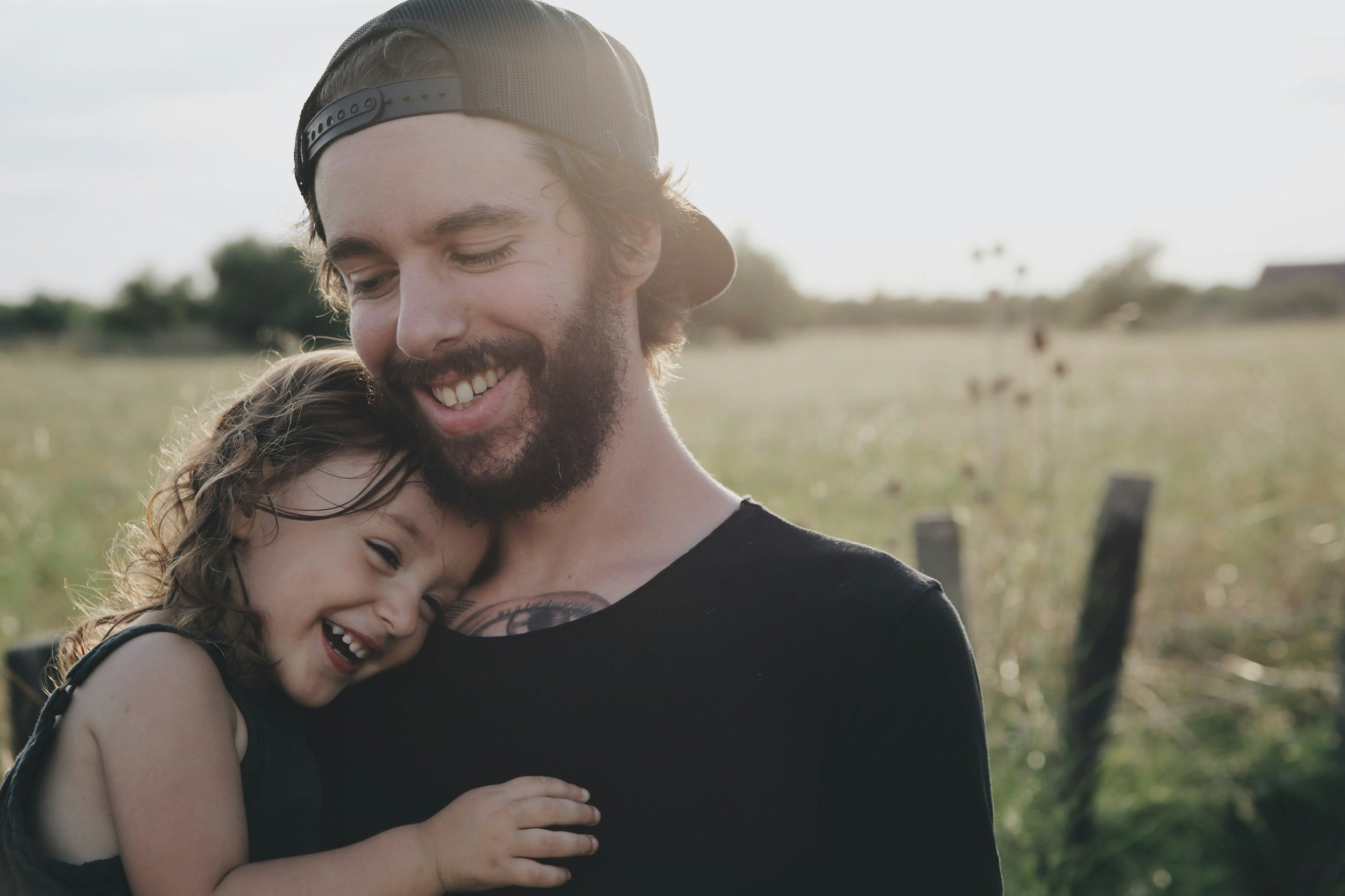 A man with a beard and wearing a backward cap is hugging a young girl in a field during the daytime, both smiling and appearing happy.