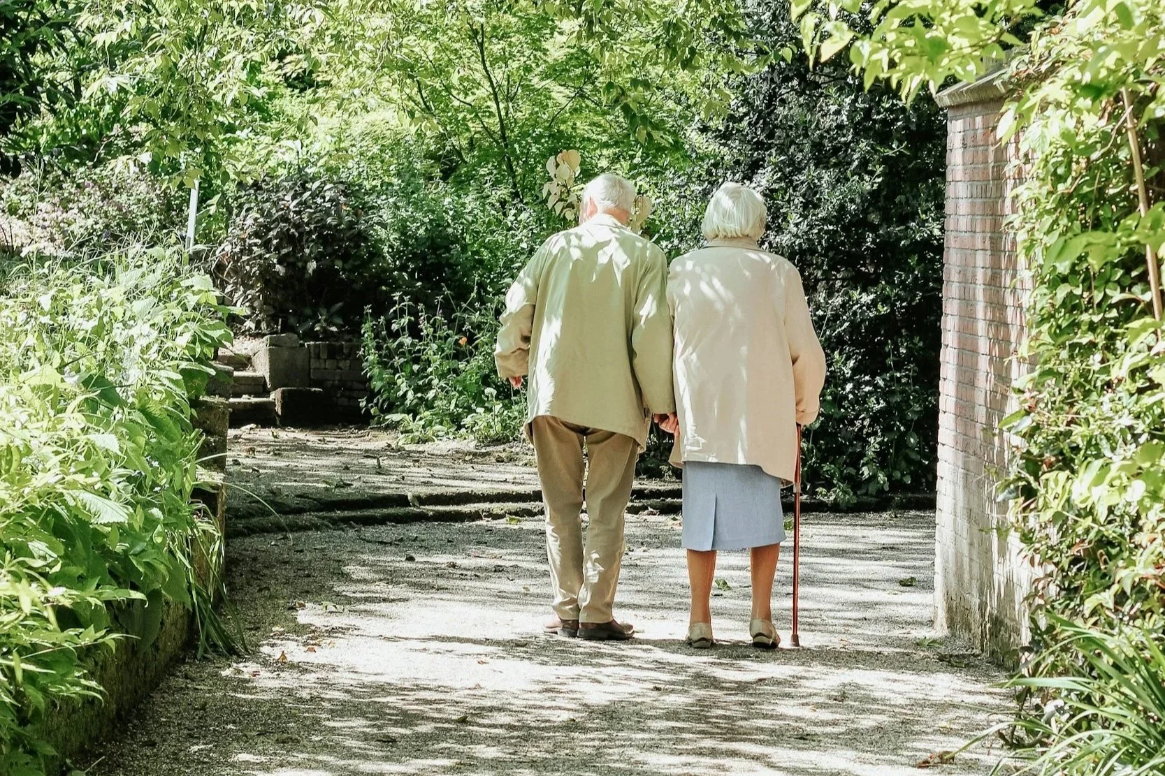 An elderly man and woman walking together on a shaded garden path with greenery and trees.