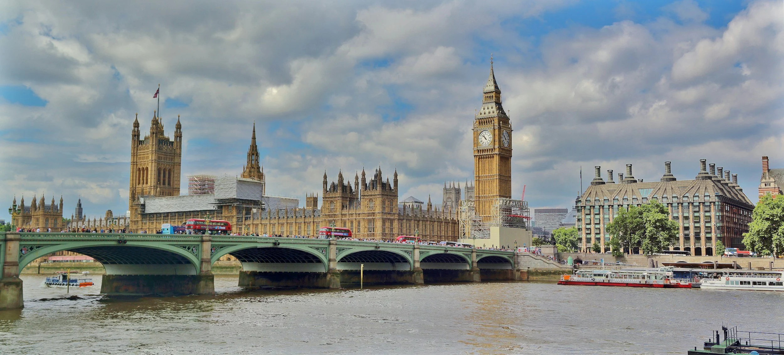 View of the Houses of Parliament and Big Ben clock tower in London, with a bridge and boats on the river Thames and partly cloudy sky.