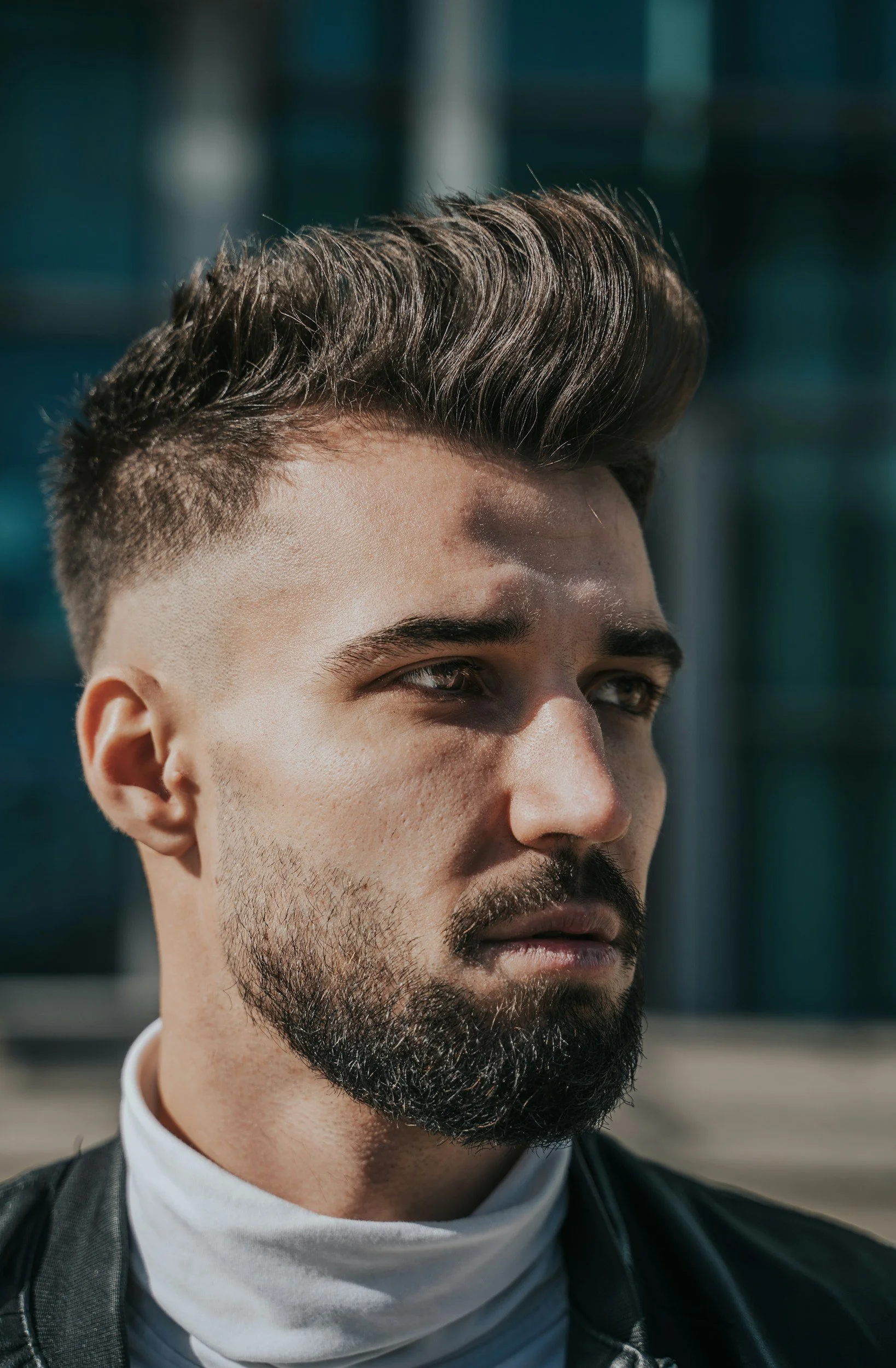 Close-up of a man with dark hair styled upward and a short beard, wearing a white turtleneck and black jacket, standing outdoors with a blurred background.