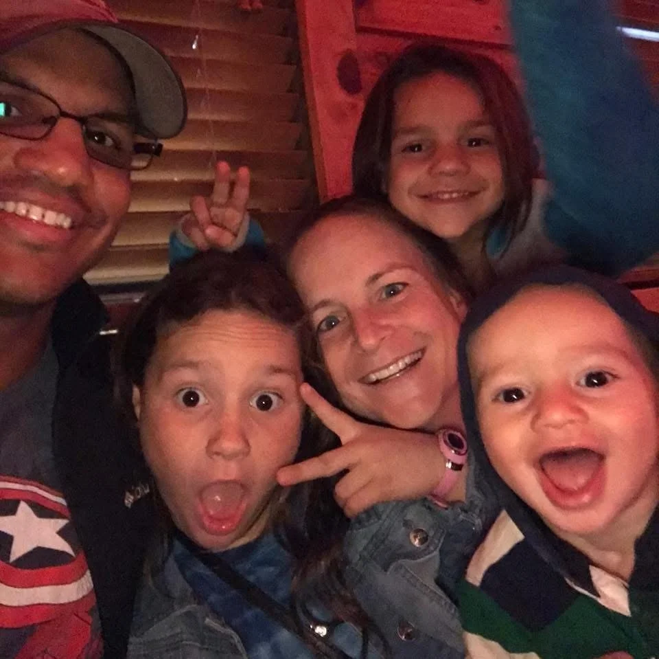 A group of six people, including two adults and four children, smiling and making playful gestures in a cozy setting with wooden decor.