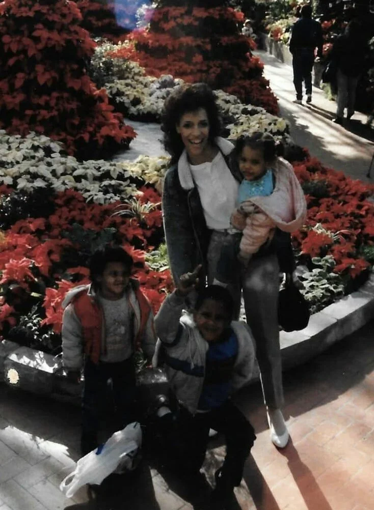 A woman and three children in a garden with overflowing red and white poinsettia flowers, some people walking in the background.