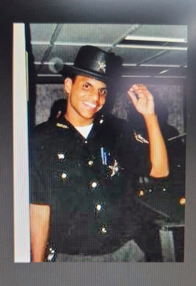 Young police officer wearing uniform and a police hat, smiling and raising his right hand in a salute or gesture of greeting, in an indoor setting.