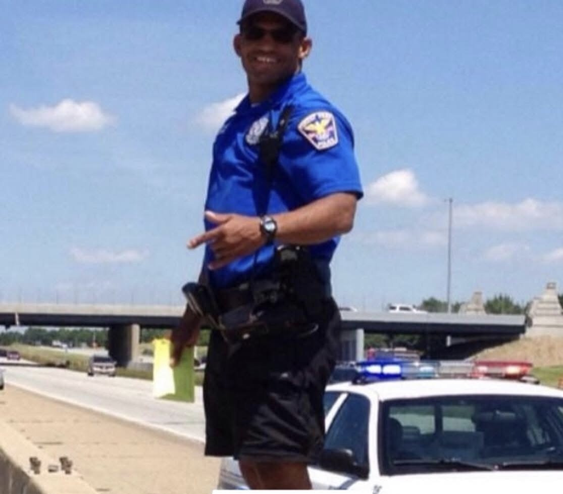 A police officer in a blue uniform stands on the side of a highway, smiling and pointing at the camera, with a police car and highway overpass in the background.