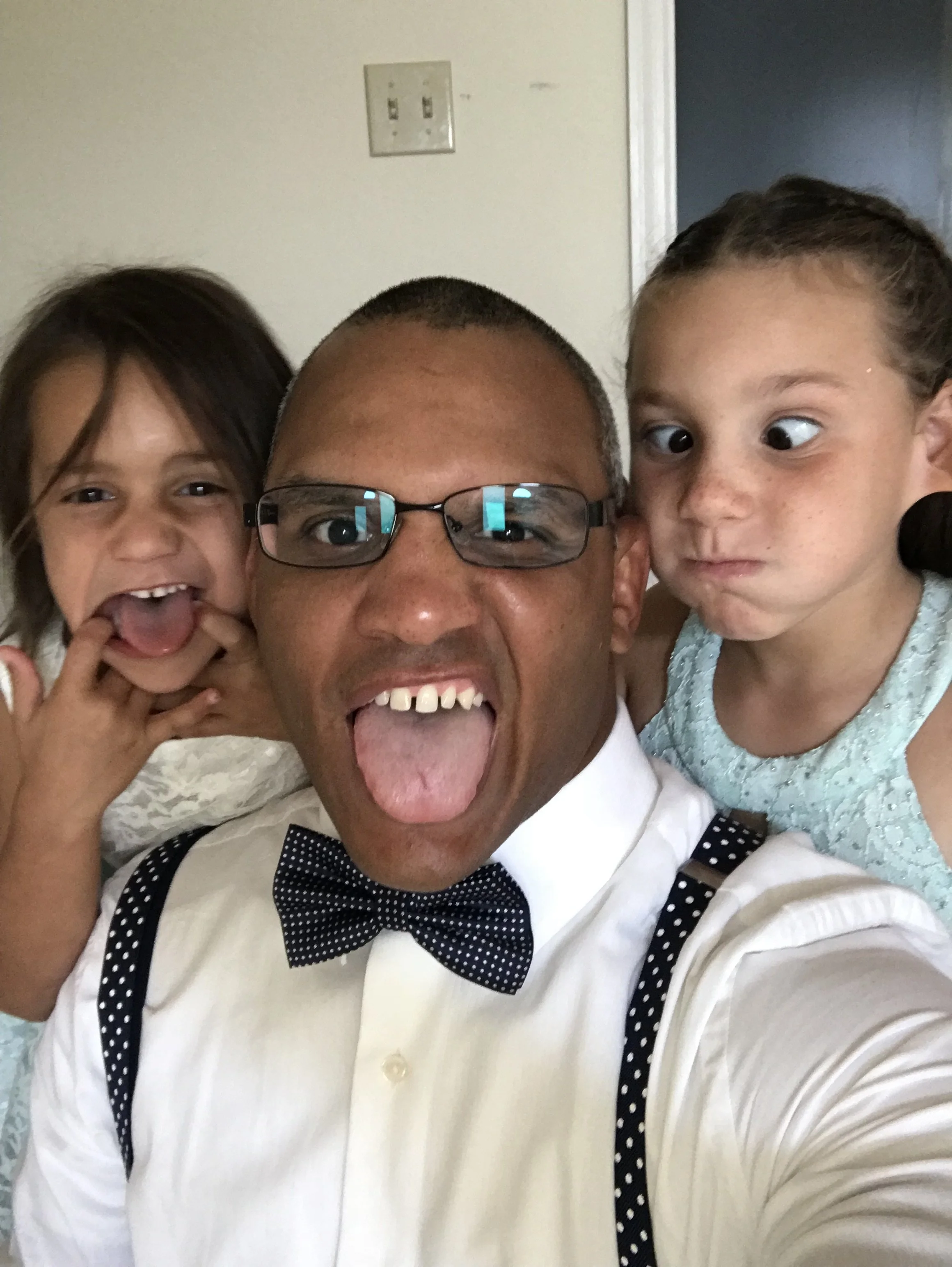 A man wearing glasses, a white shirt, and a polka dot bow tie is taking a selfie with two young girls. The man and one girl are sticking out their tongues, and the other girl is making a funny face with wide eyes and pursed lips.