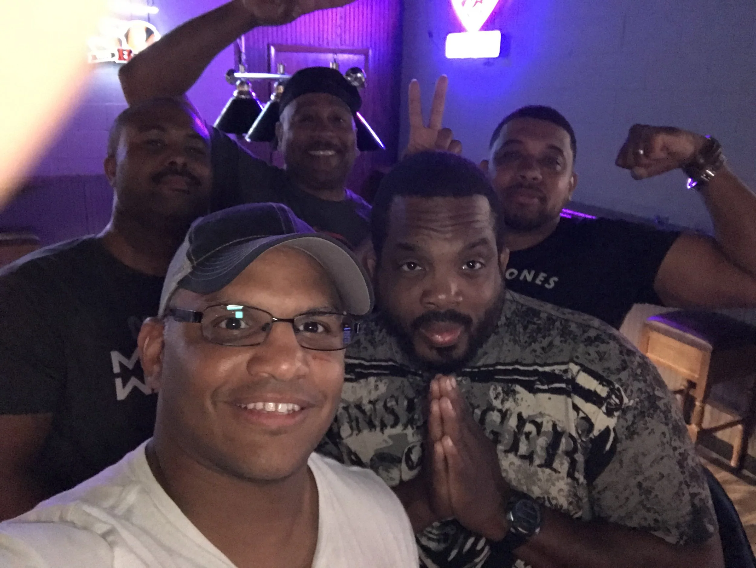Group of five men taking a selfie inside a dimly lit bar or restaurant with purple lighting and neon signs in the background.