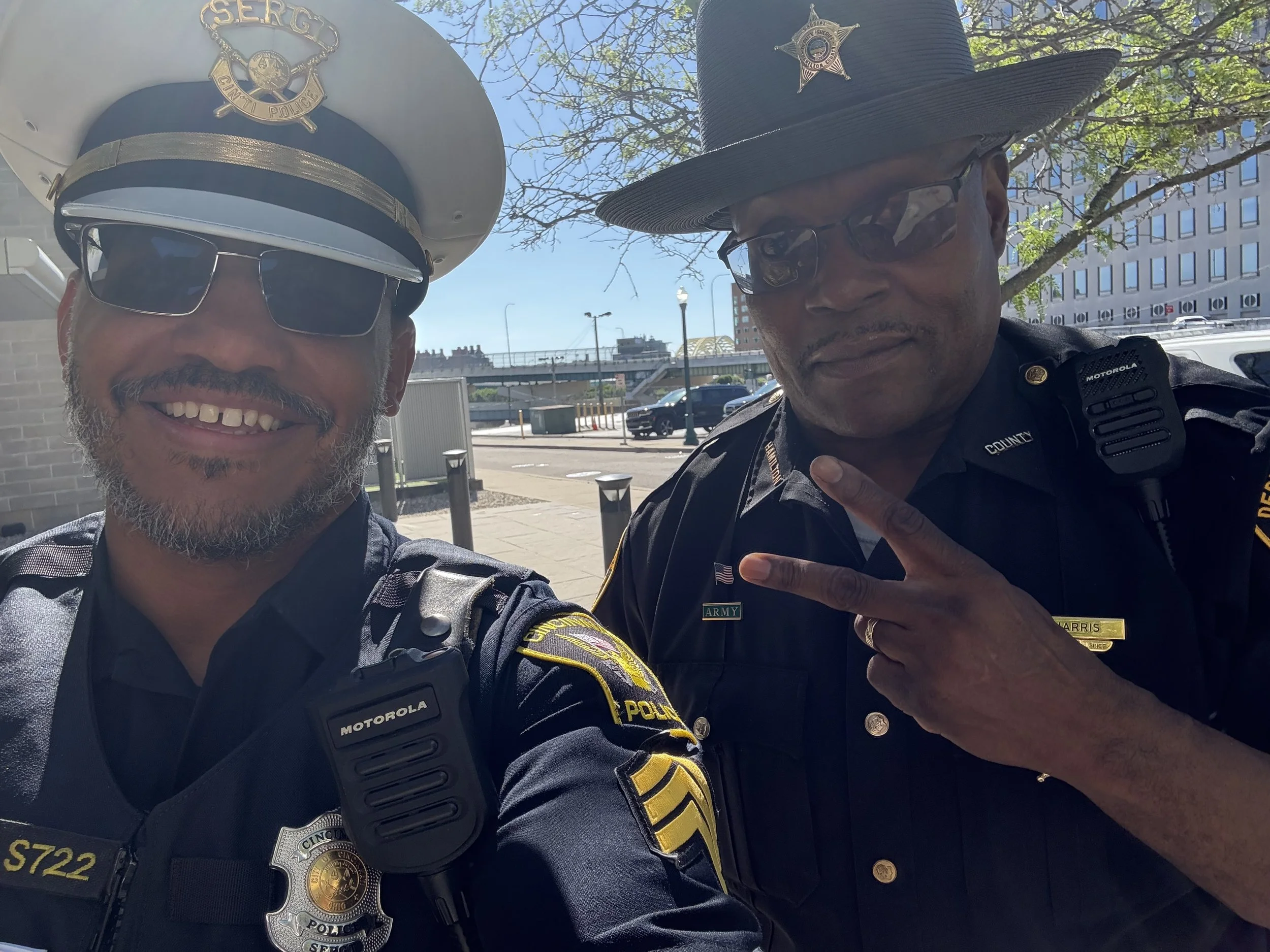 Two police officers taking a selfie outdoors on a sunny day, wearing police uniforms and hats, with city buildings and a street in the background.