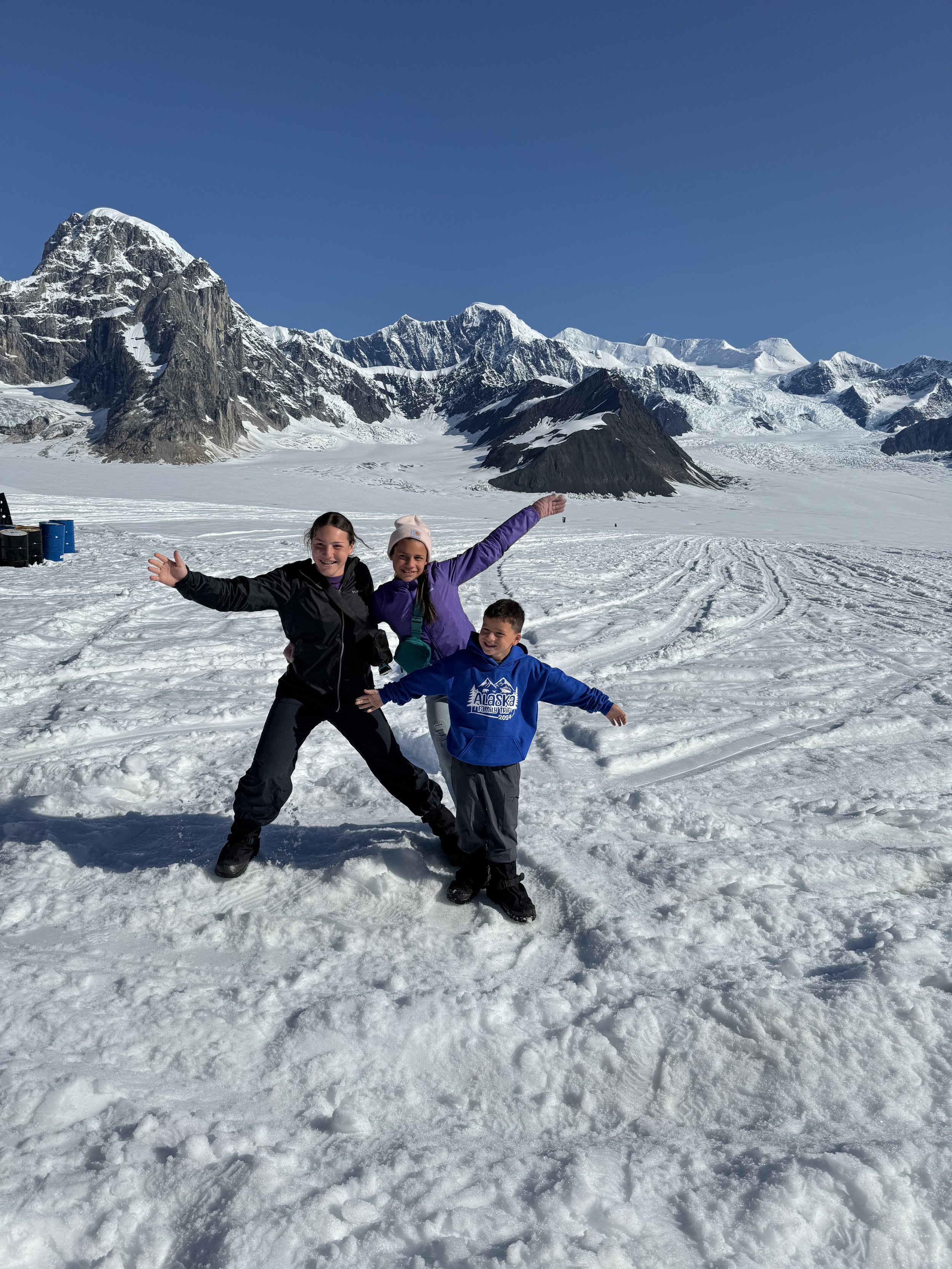 Three children posing and having fun in a snowy landscape with mountains and glaciers in the background, wearing winter jackets and boots. Alaska. Ruth Glacier.