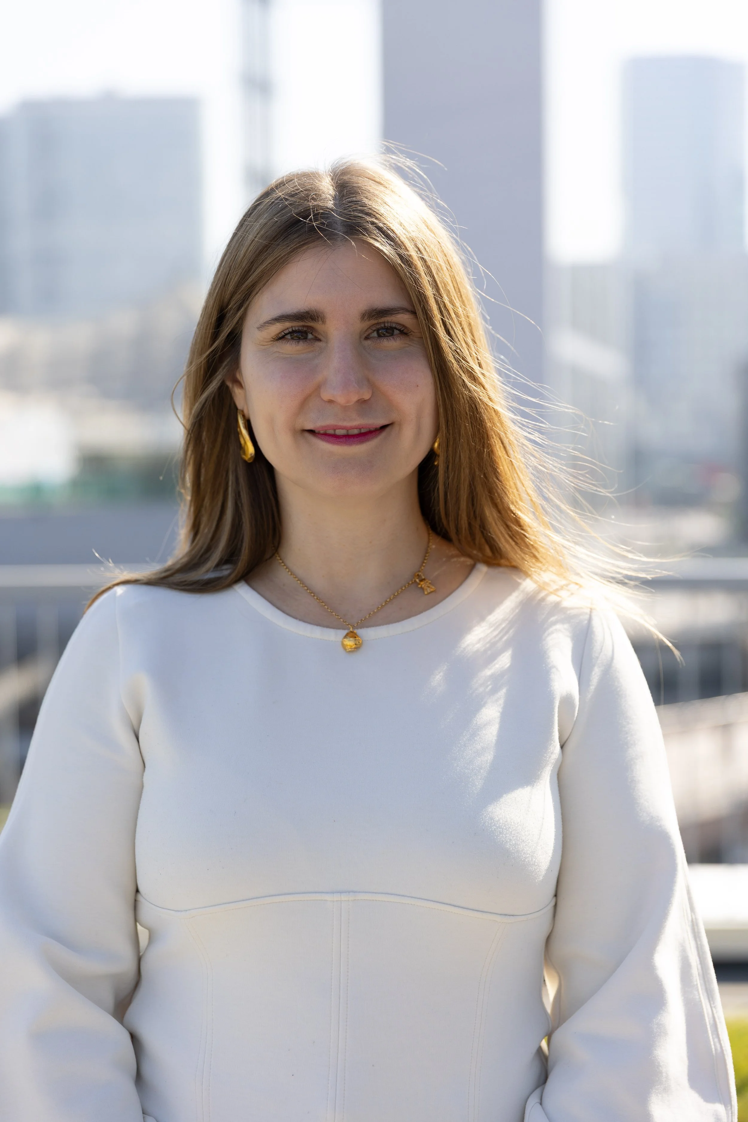 Professor Maja Koricar smiling outdoors in front of a cityscape with tall buildings, wearing a white top and gold jewelry.