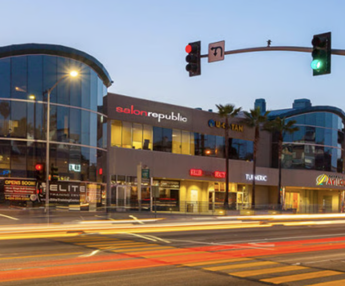 Commercial building with stores, including Salon Republic and Quick Tan, on a city street at dusk, with traffic lights and light trails from passing cars. Med Spa in Hermosa Beach, CA | Nua Aesthetics
