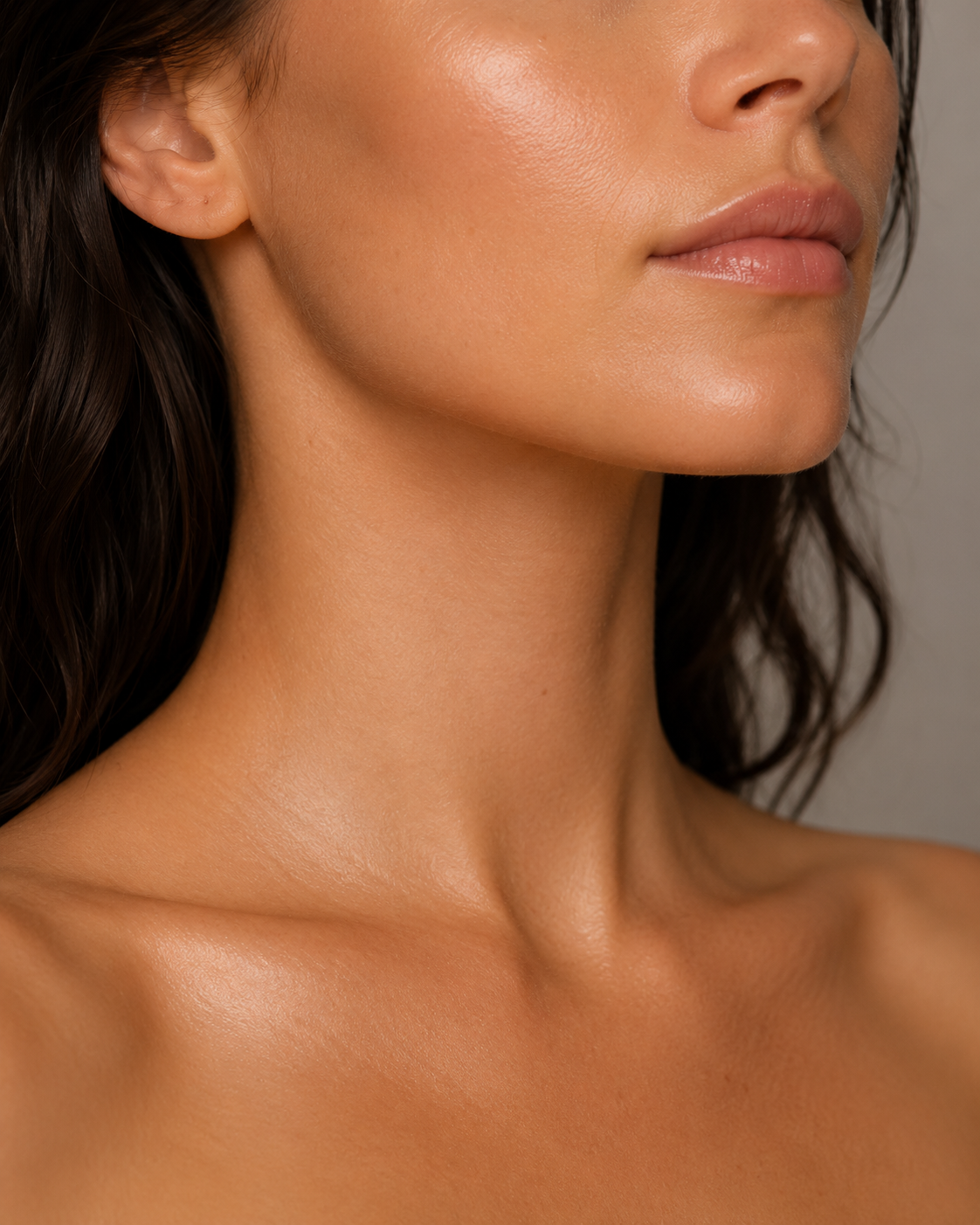 Close-up of a woman's lower face, neck, and shoulder, with smooth, glowing skin, dark wavy hair, and minimal makeup.