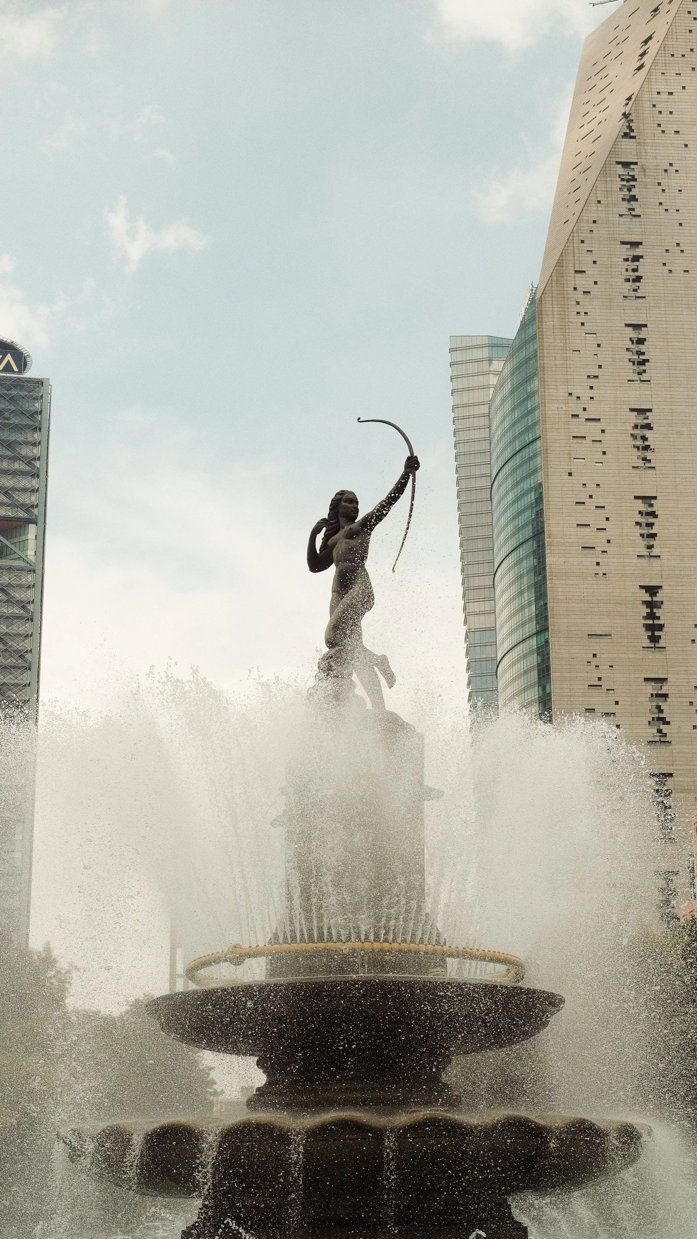 Fuente con escultura de una mujer con arco en la parte superior, en un entorno urbano con edificios modernos y cielo nublado.