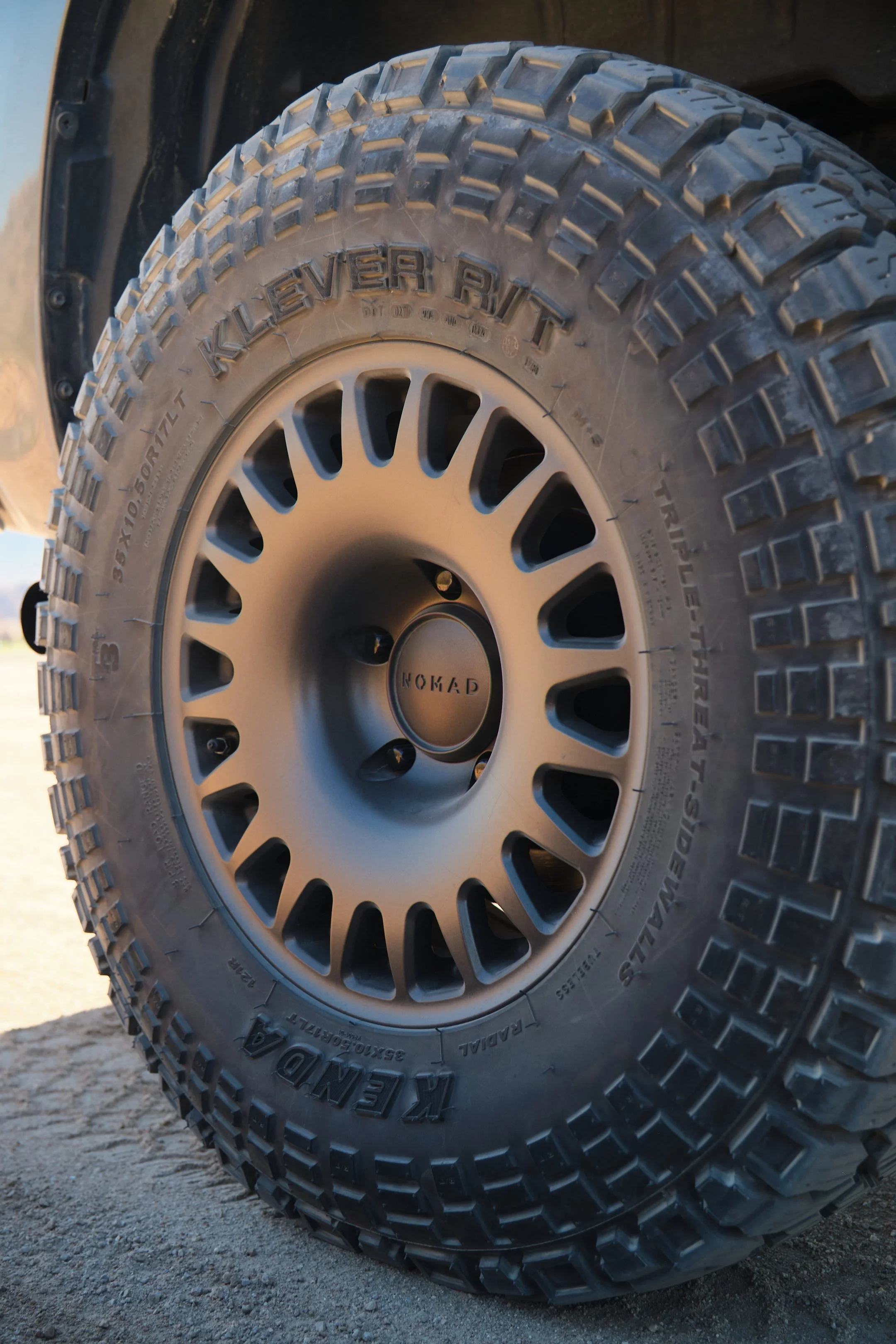 Close-up of a rugged off-road tire mounted on a black rim with the 'NOMAD' logo at the center, on a dirt surface.