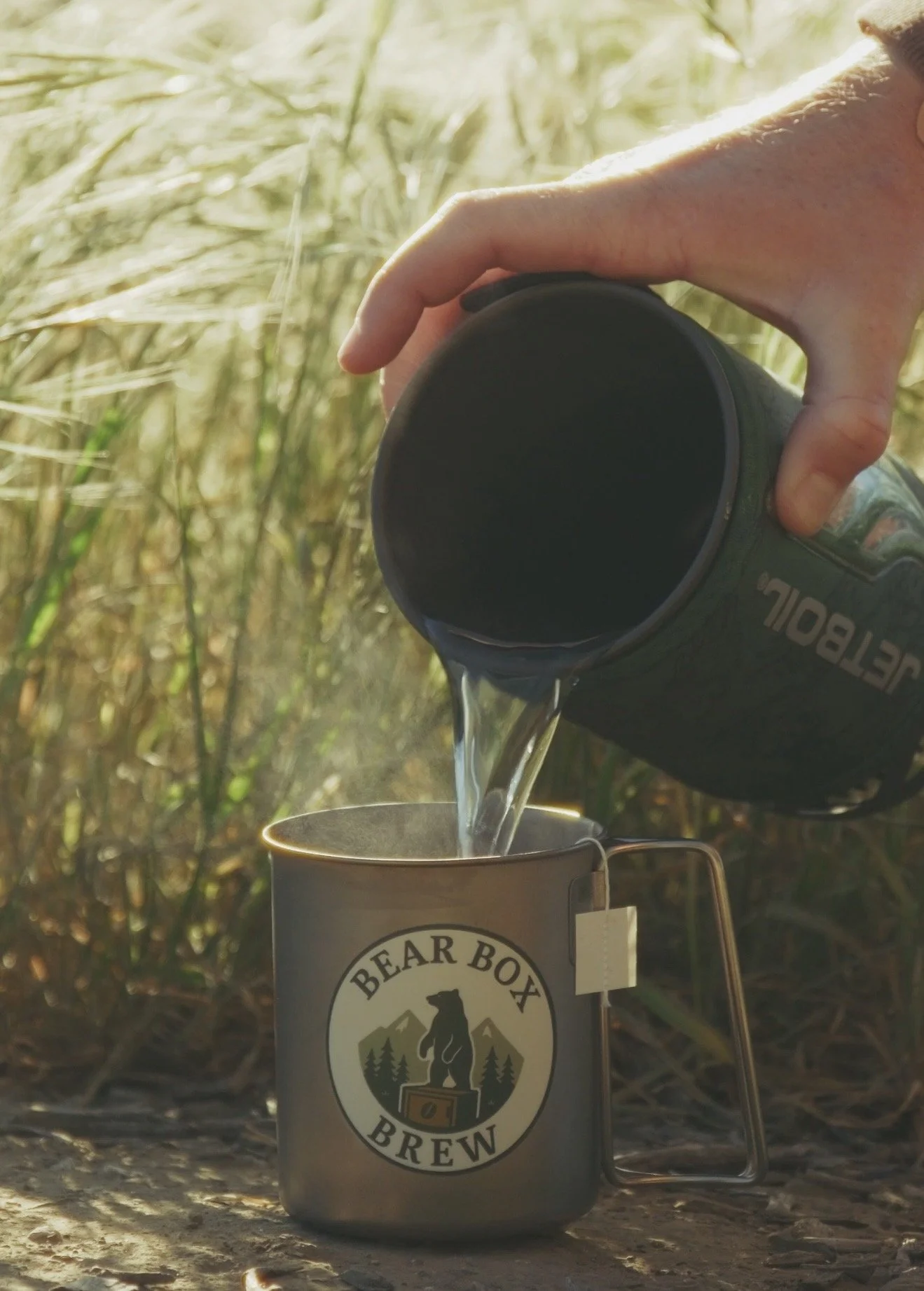 A person pours hot water into a mug with a Bear Box Brew logo outdoors in a grassy area.
