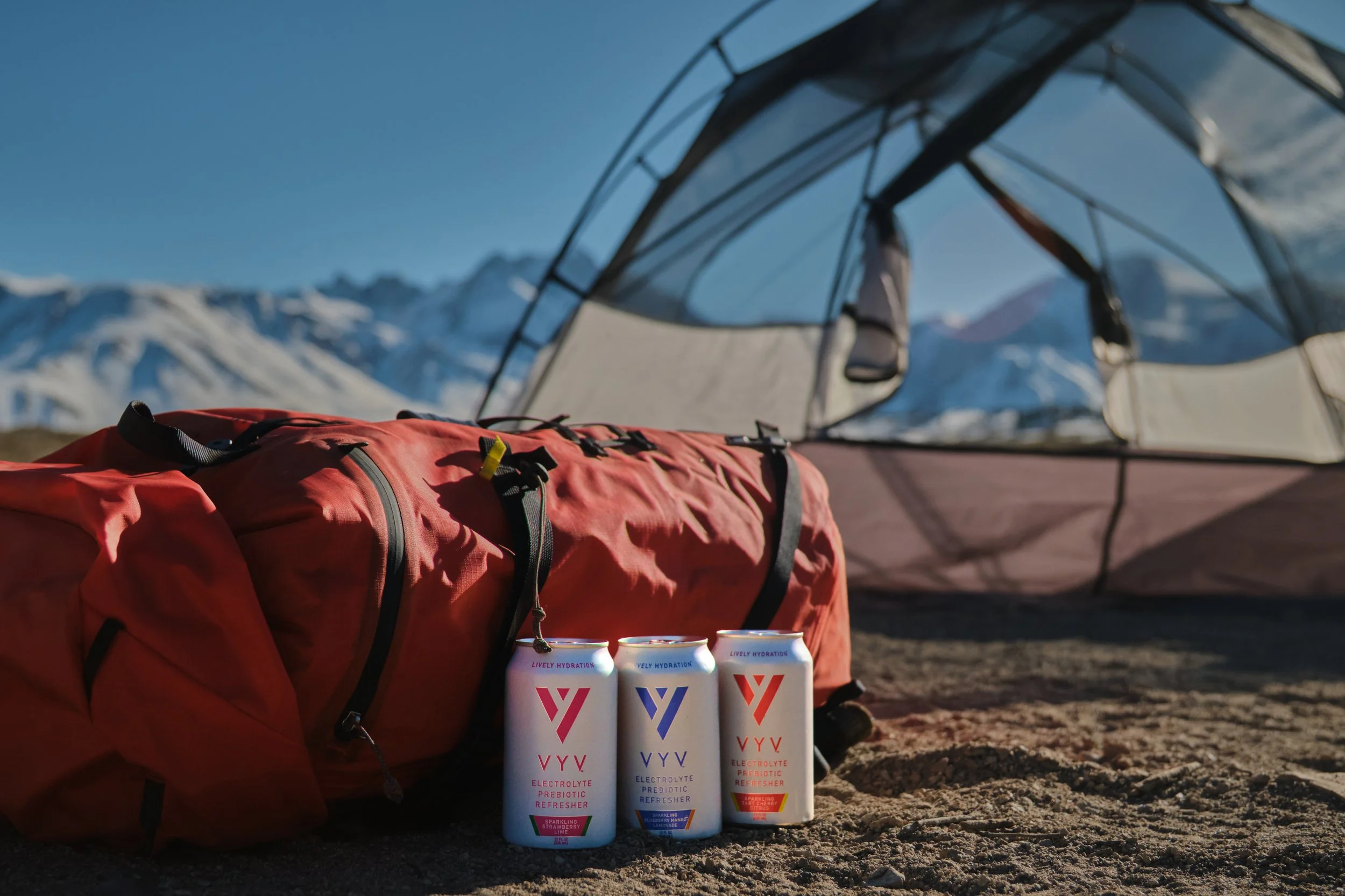 Camping scene with a large red backpack, three cans of VVV electrolyte prebiotic refresher, and a tent set up on rocky ground with snow-capped mountains in the background.