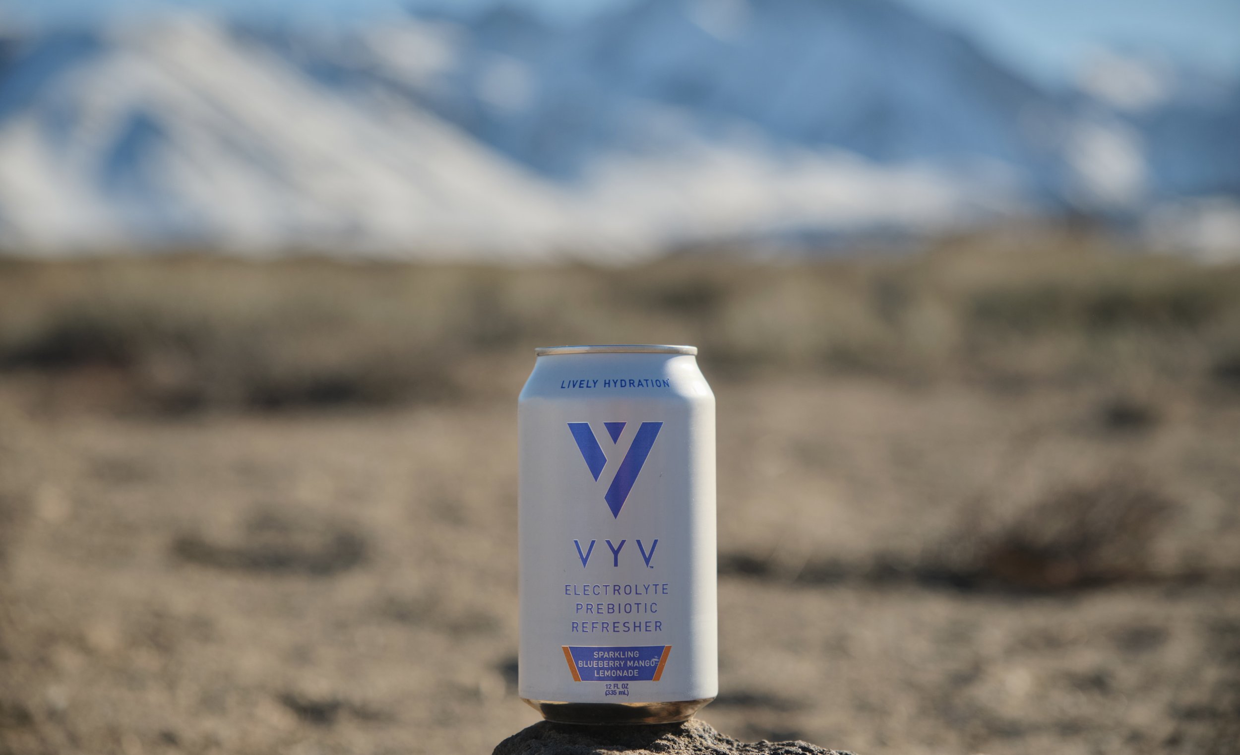 A can of VYV electrolyte prebiotic refresher with blueberry mango lemonade flavor placed on a rock in a desert with mountains and sky in the background.