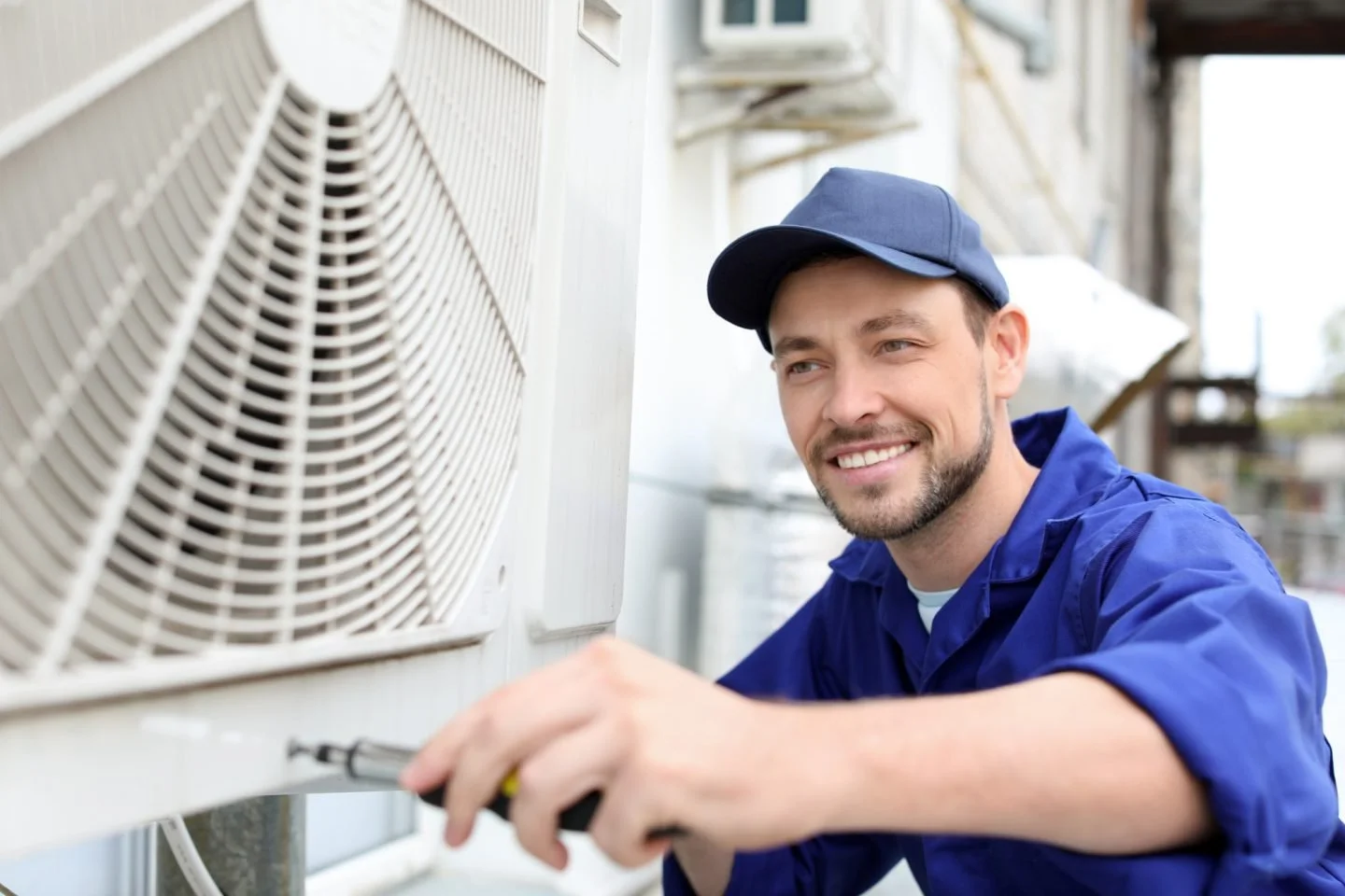A smiling technician in a blue uniform and cap using a screwdriver on an outdoor HVAC unit.