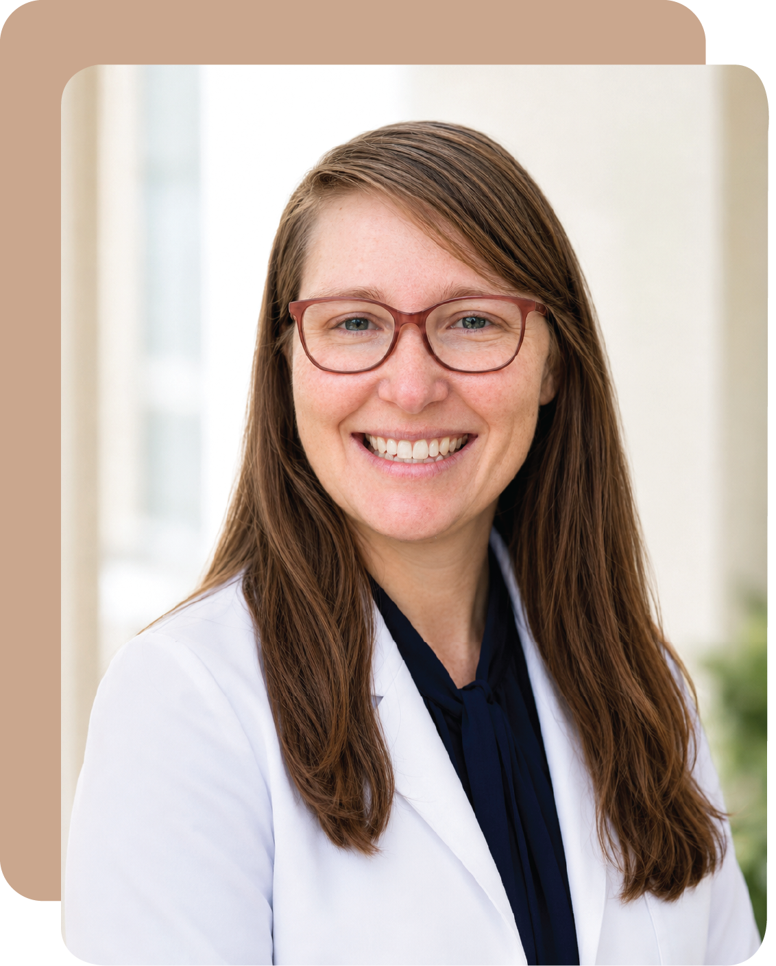 A woman with long brown hair, wearing glasses and a white lab coat, smiling indoors with a blurred background.
