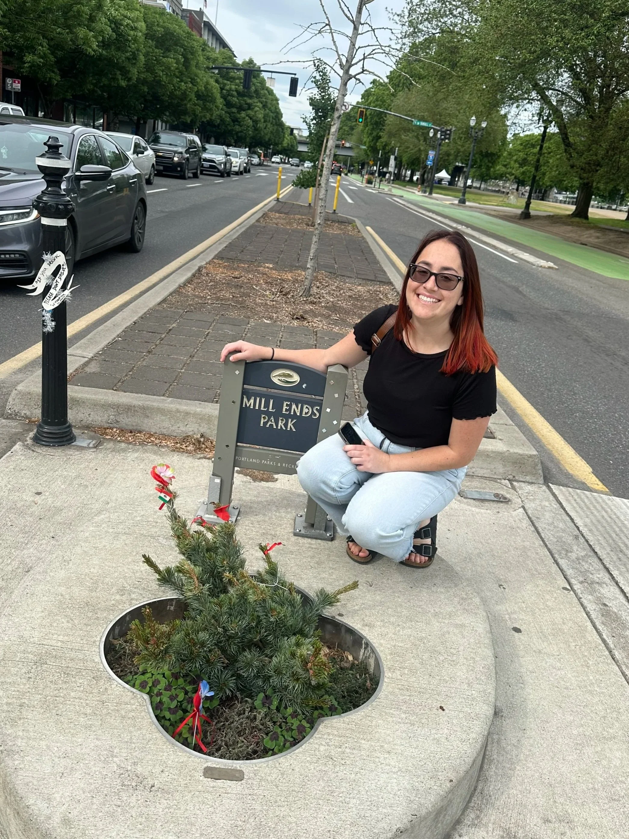 A young woman with red hair and glasses smiling at the camera while kneeling next to a sign that reads 'Mill Ends Park' in Portland, Oregon. She is holding a phone in her right hand. There are small memorial flowers and a miniature Christmas tree with ribbons in the flower bed in front of her. The background shows a street with parked cars, green trees, and city infrastructure.