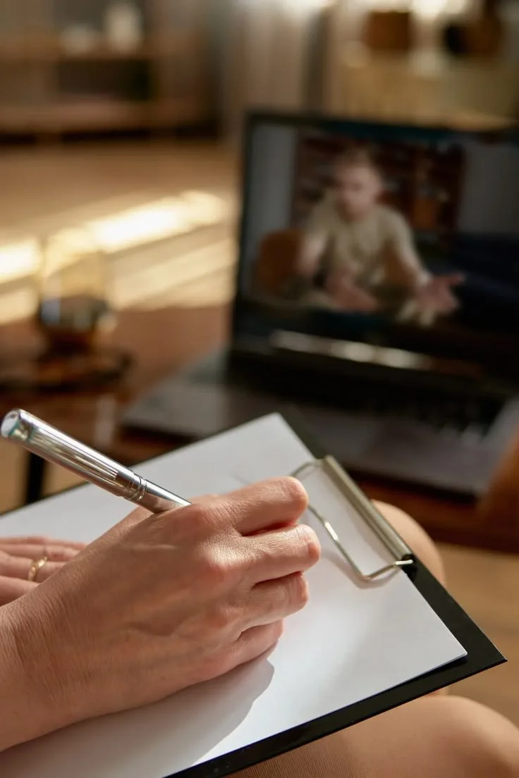A person taking notes on a clipboard with a silver pen during a video call on a laptop, with a blurred background showing a room.