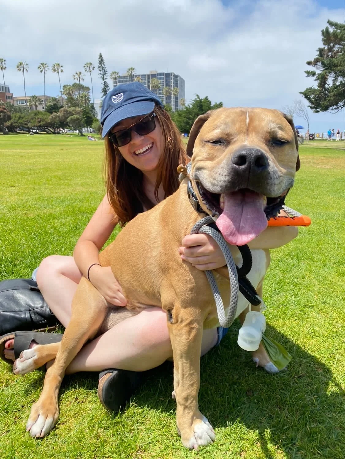 A woman with long red hair wearing sunglasses (Yana Levin) , a blue cap, and a black tank top, smiling while sitting on grass at a park with her large, happy tan-colored dog.