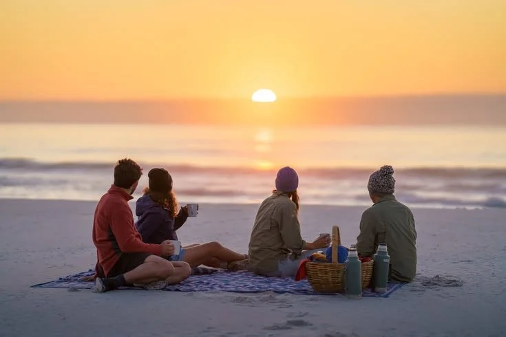 Four people sitting on a blanket at the beach during sunset, having a picnic with drinks and snacks.