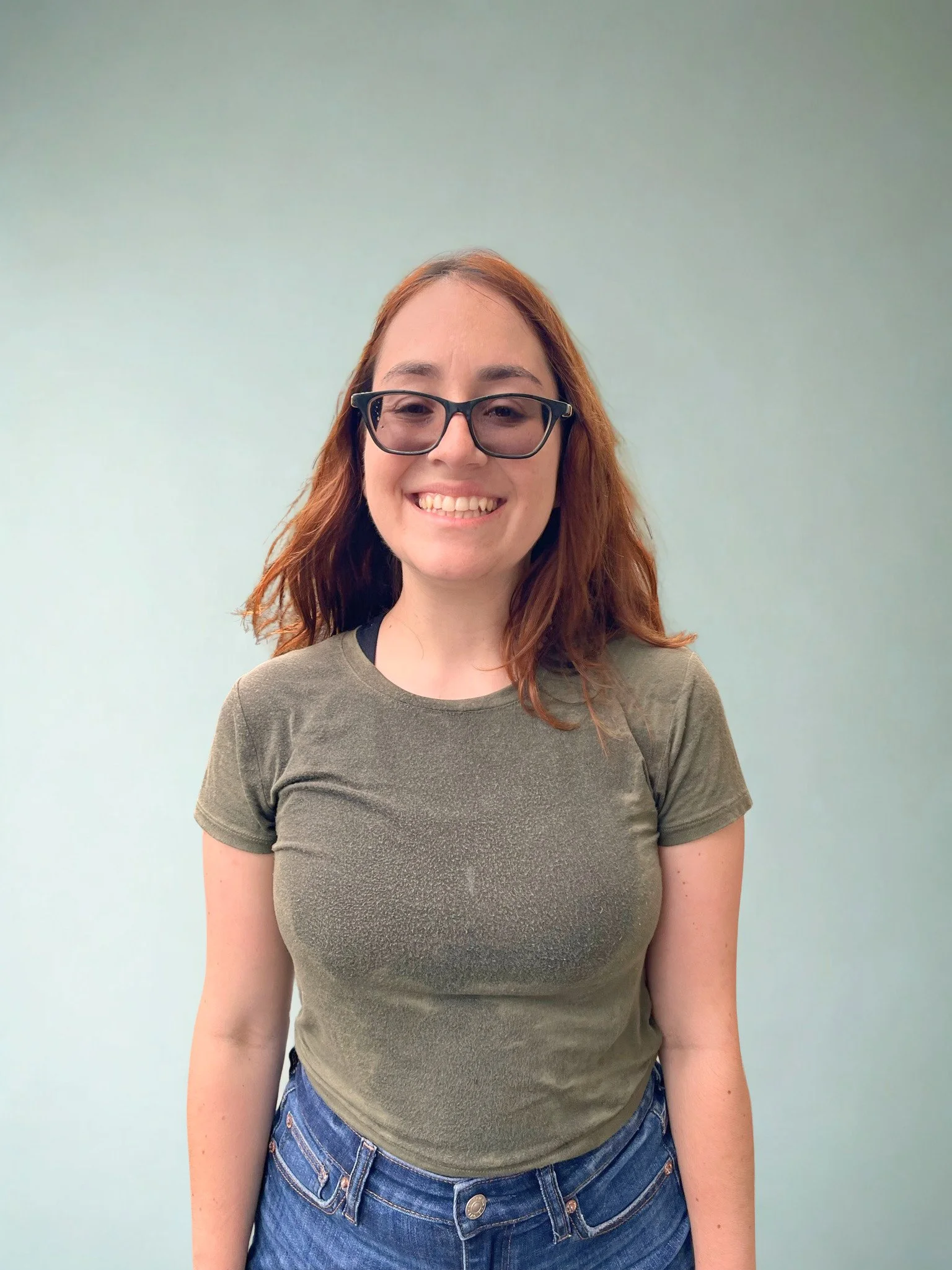 A young woman with red hair, wearing glasses, a green t-shirt, and blue jeans, smiling in front of a plain light green wall.
