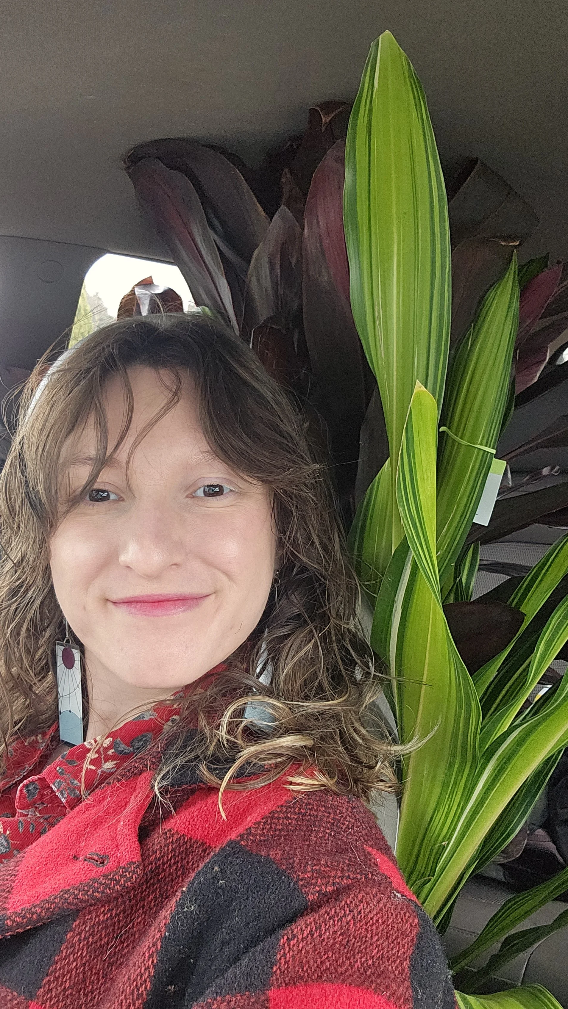 Close-up of a smiling woman with curly brown hair, wearing a red and black checkered jacket, standing next to tall green plants inside a vehicle