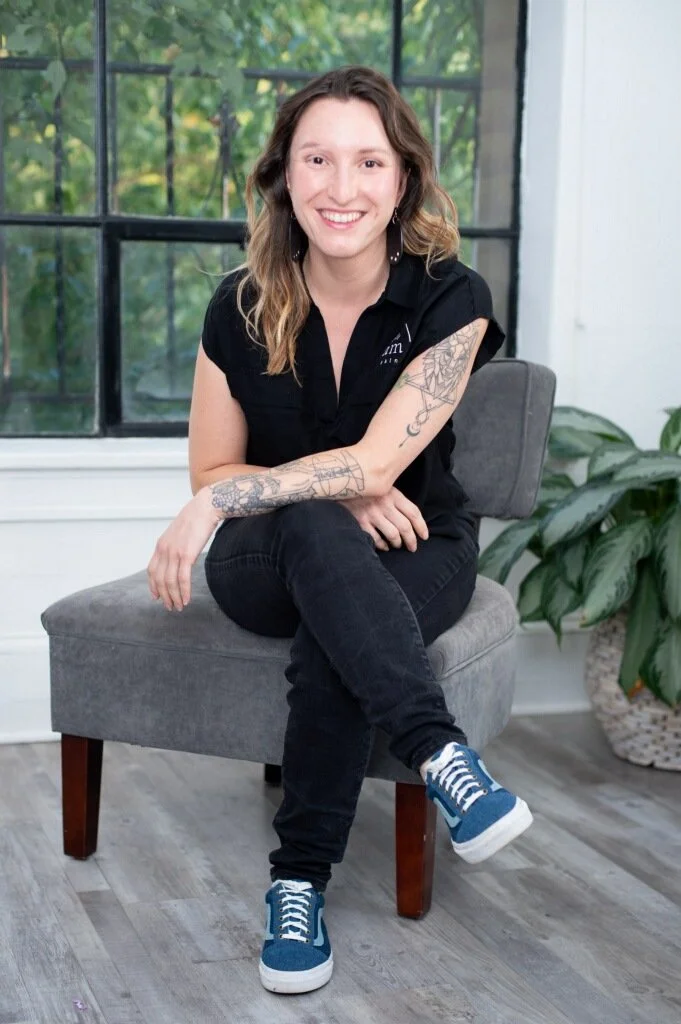 A smiling woman with wavy hair, wearing a black shirt with a logo, dark jeans, and blue sneakers, sitting on a gray chair in a room with a large window and green plants.