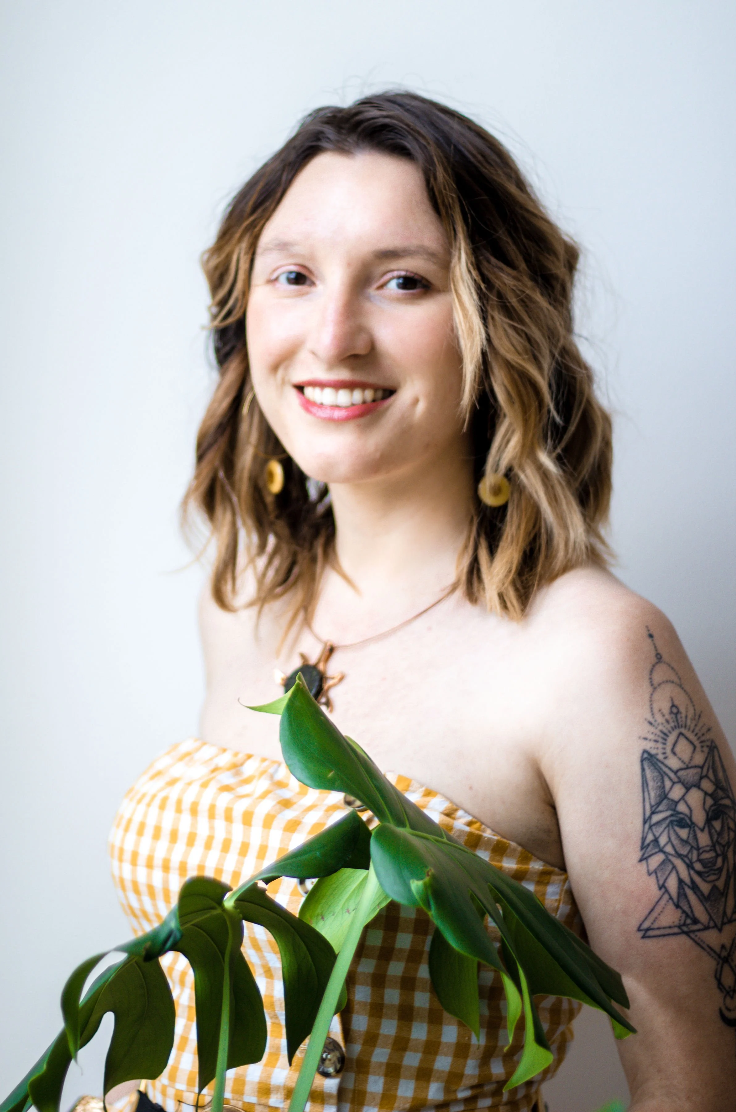 Smiling woman with shoulder-length wavy brown hair and tattoos on her right arm, wearing a yellow checkered off-shoulder top, holding a large green plant with broad leaves, standing against a plain light-colored background.