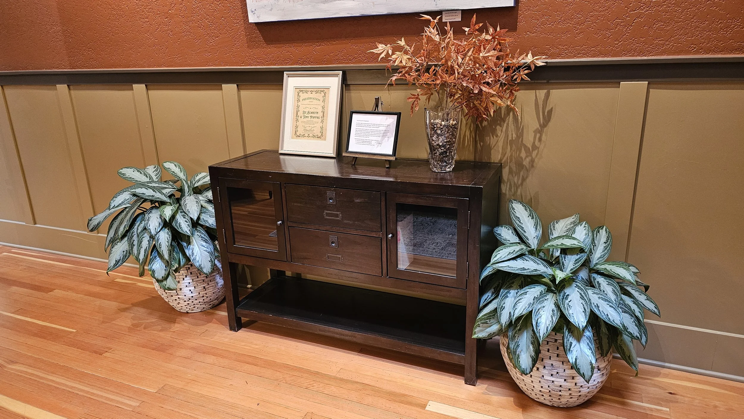 A dark wooden cabinet with glass doors and open shelving, decorated with framed certificates and a glass vase filled with orange and red leaves, flanked by two potted variegated foliage plants in woven baskets, against a brown and beige wall with woo