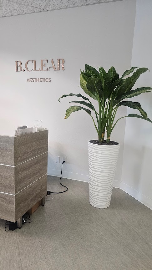 Interior of a clinic or aesthetic office with a large green houseplant in a white textured planter and a reception desk against a white wall with a sign that reads 'B. CLEAR AESTHETICS'.