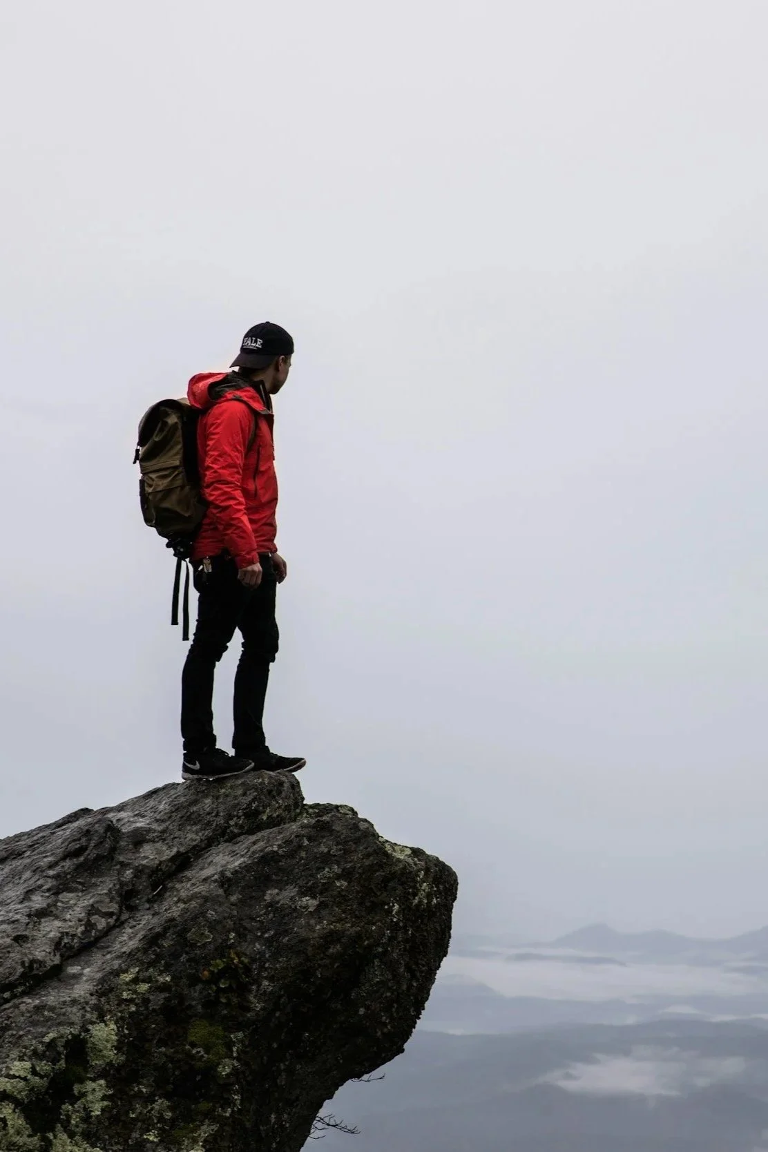 photograph of a hiking man in a red jacket and backpack on top of a mountain looking out to a mountain range viewpoint