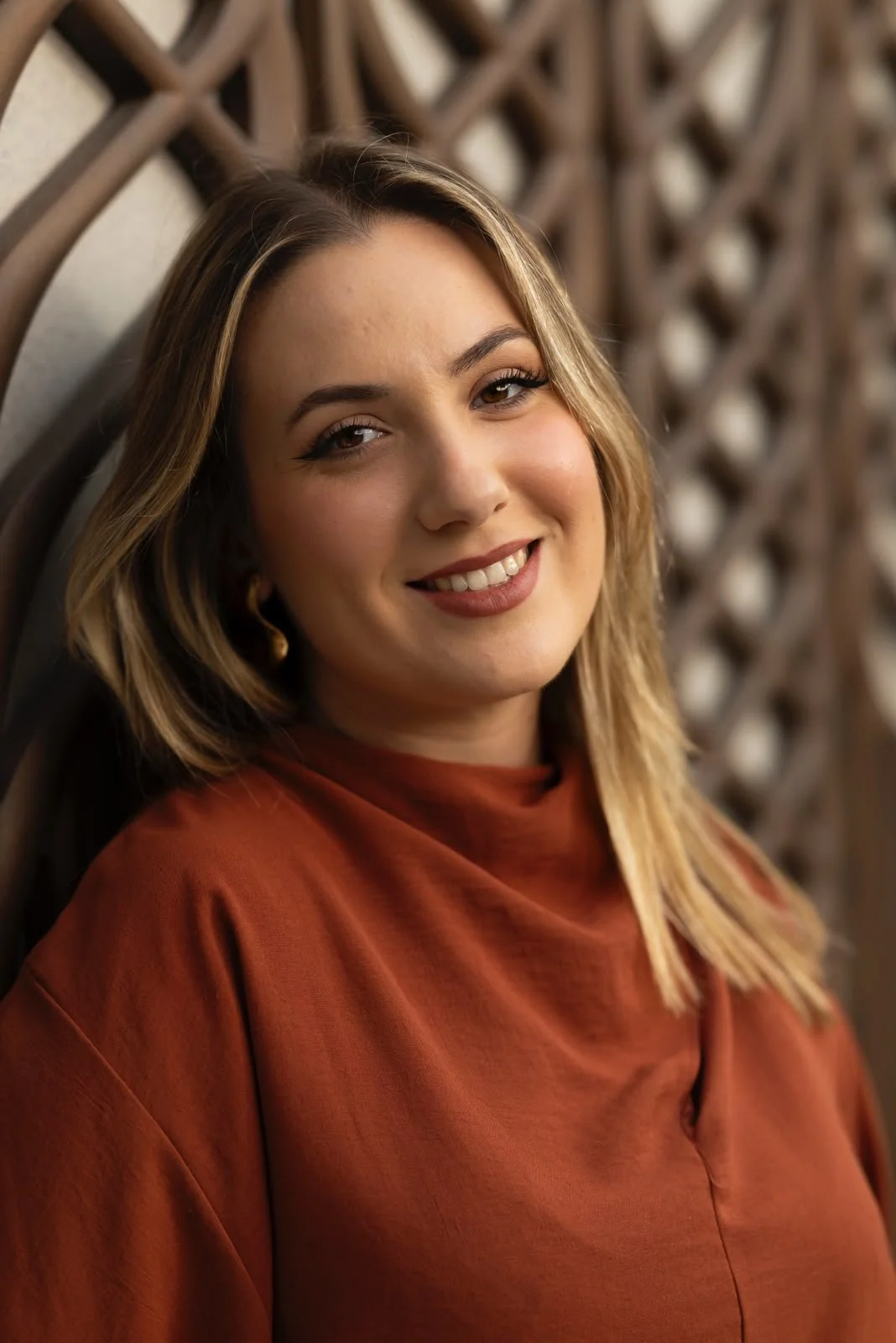 Retrato de una mujer con cabello rubio, sonrisa y ropa naranja, posando contra una cerca de madera en un ambiente cálido.