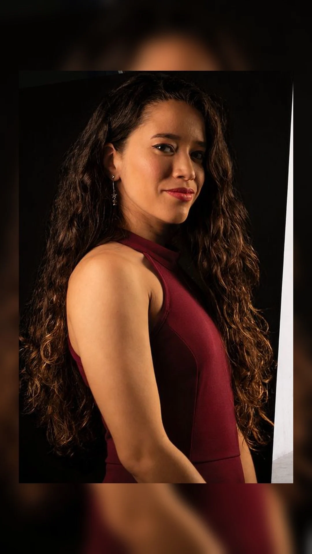 Retrato de una mujer con cabello largo y rizado, usando un vestido sin mangas de color burdeos, contra fondo negro.