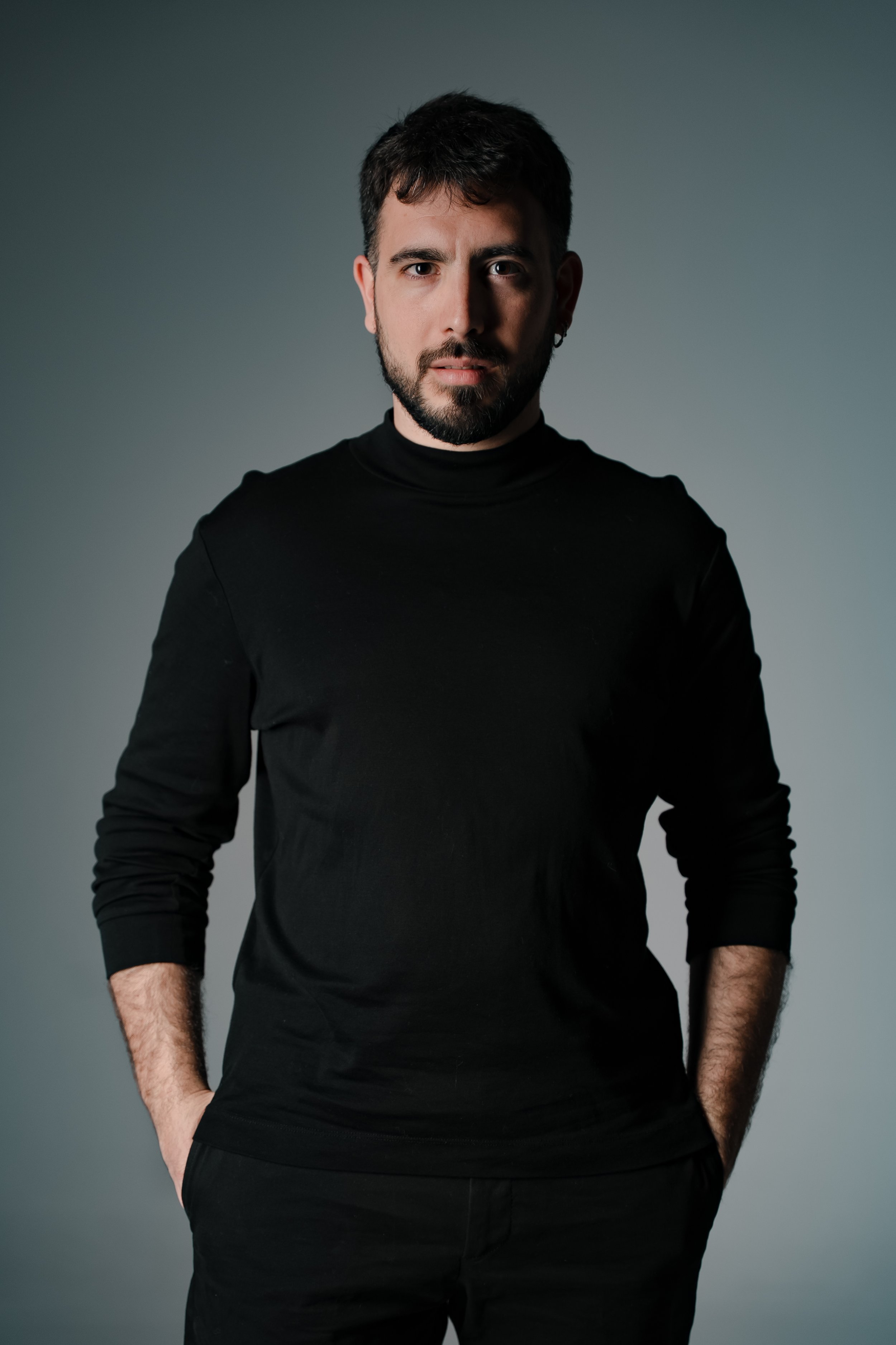 Retrato de un hombre con barba y cabello oscuro, vistiendo una camiseta negra, en un fondo gris.