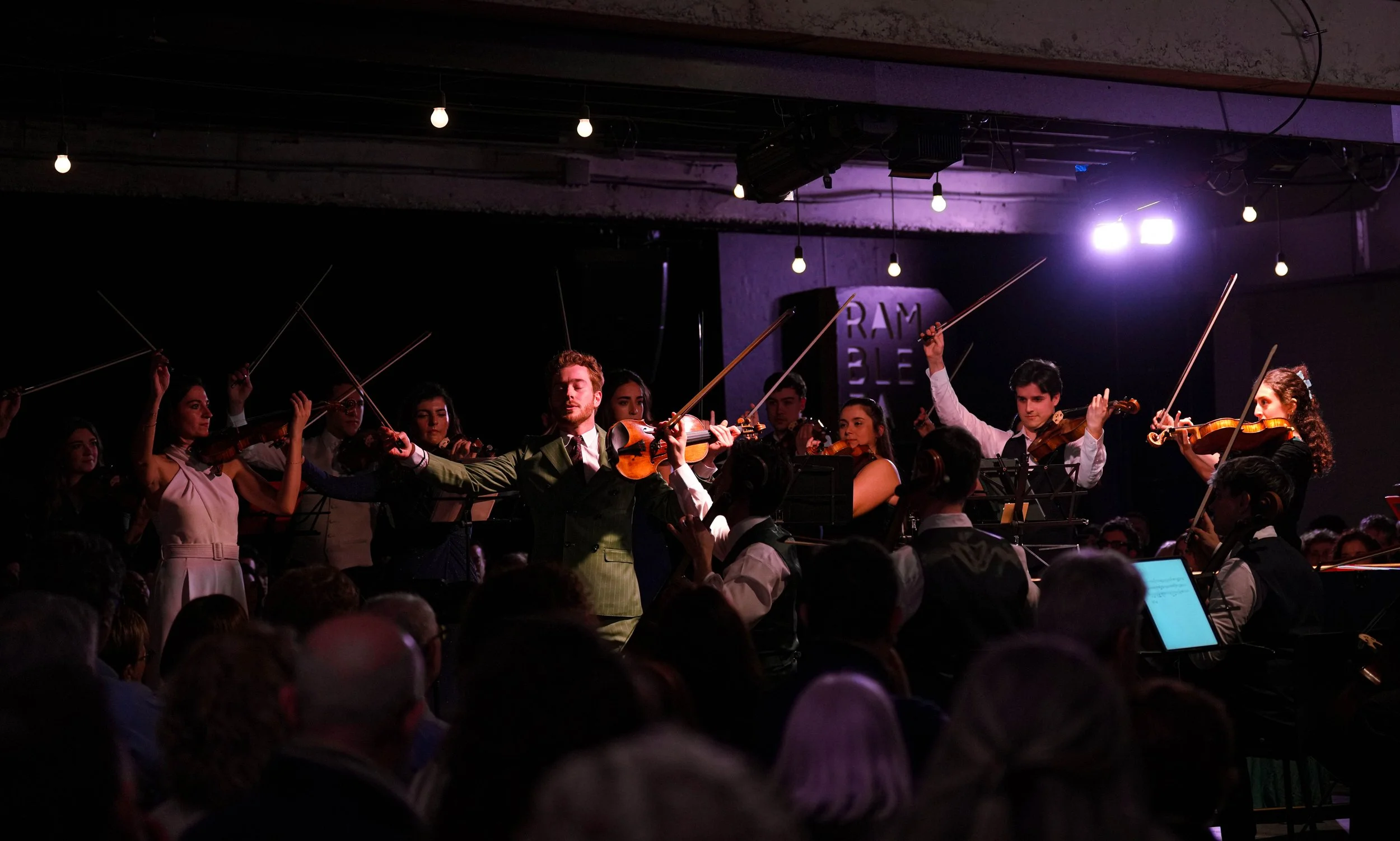 Orquesta de jóvenes tocando violines en un escenario con luces violetas y fondo oscuro, audiencia mirando.