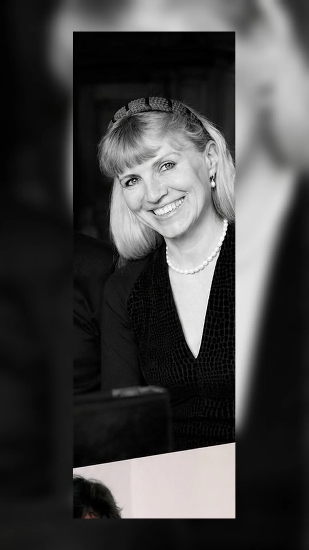 Retrato en blanco y negro de una mujer sonriendo, con cabello rubio, usando un collar de perlas y pendientes, vestida con blusa negra.