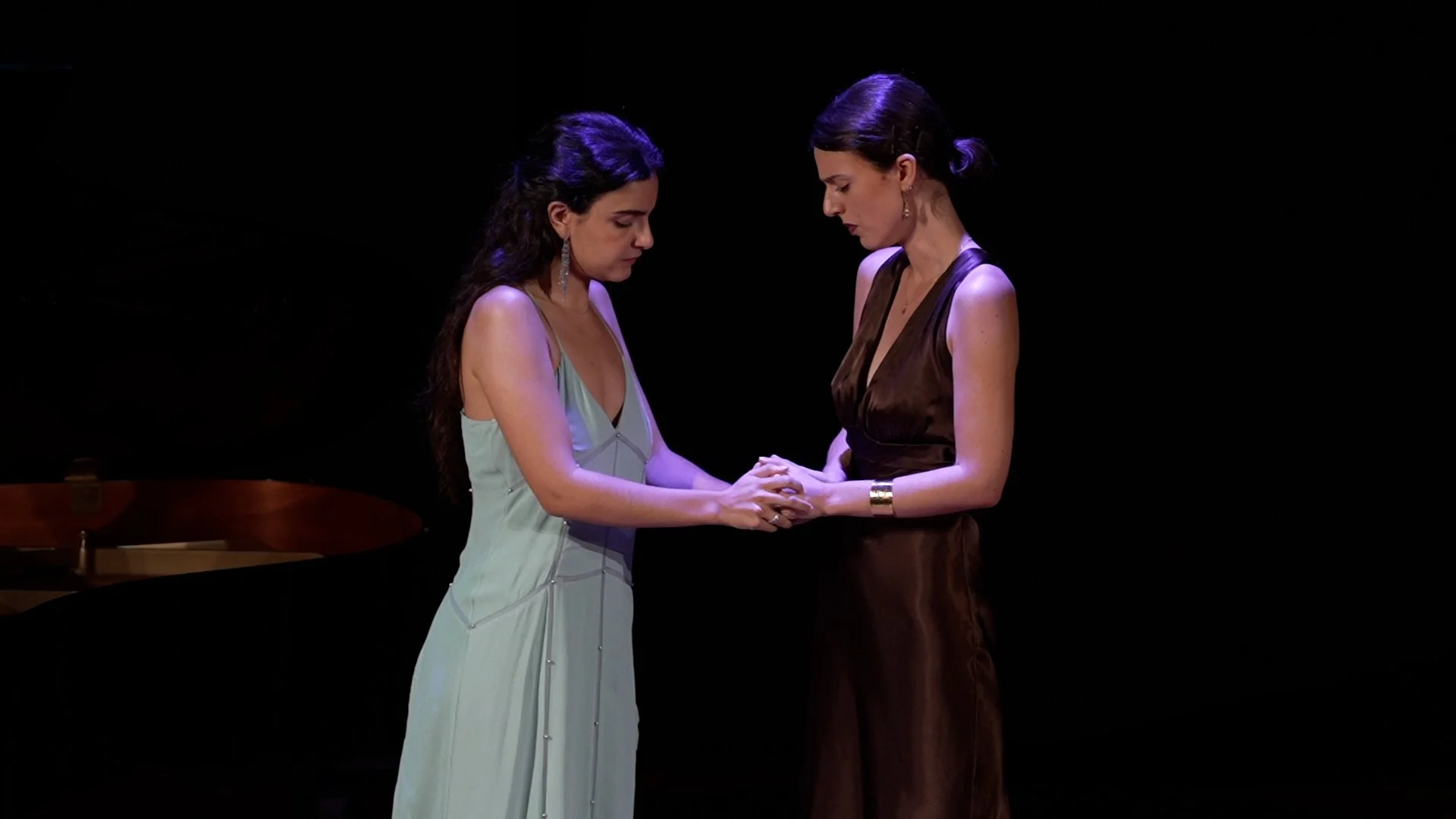 Dos mujeres con vestidos elegantes sosteniéndose las manos en un escenario con fondo negro.