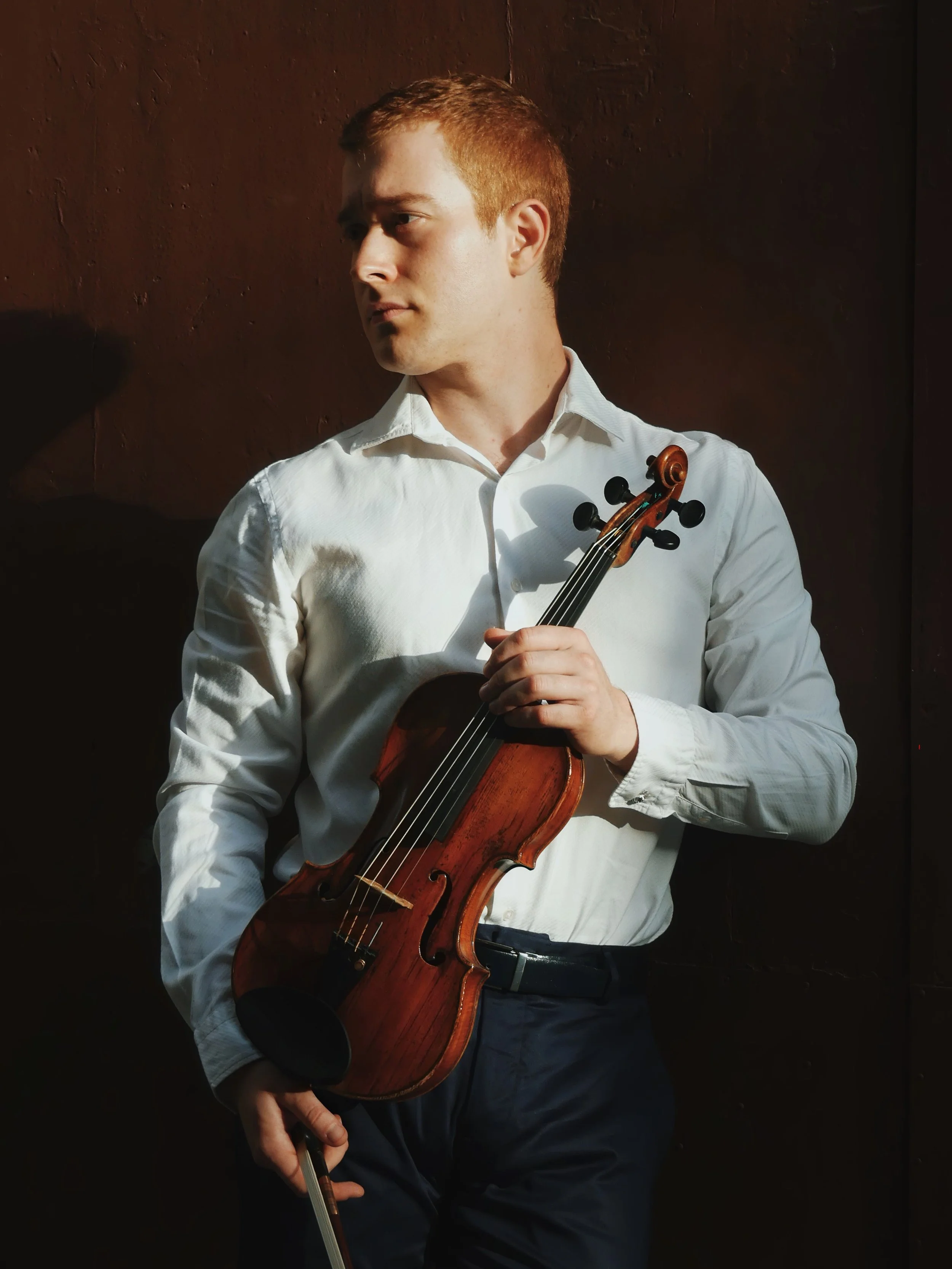 Joven con camisa blanca sostiene un violín y un arco, posando frente a una pared oscura.