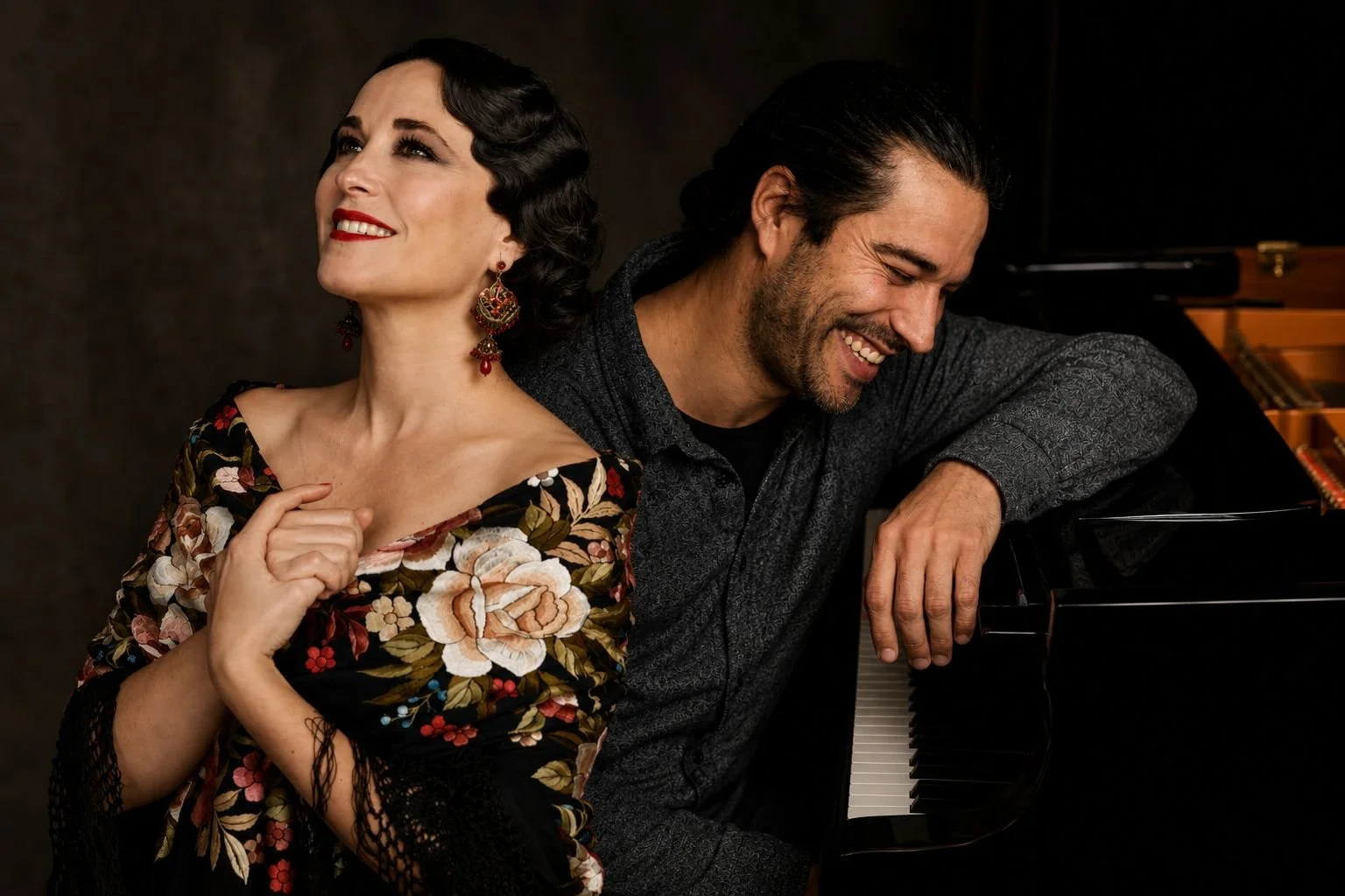 Una pareja sonriente, una mujer con vestido floral y pendientes largos, y un hombre con camisa gris oscuro, junto a un piano.