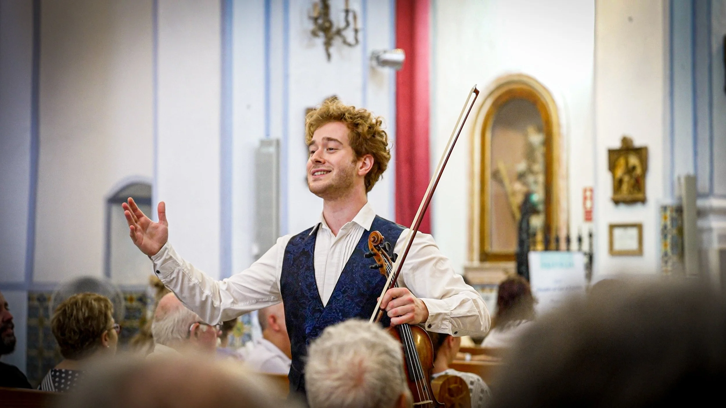 Hombre tocando el violín en un concierto, aparentemente dirigiendo la orquesta, en una iglesia o sala de eventos decorada con cuadros y detalles artificiales.